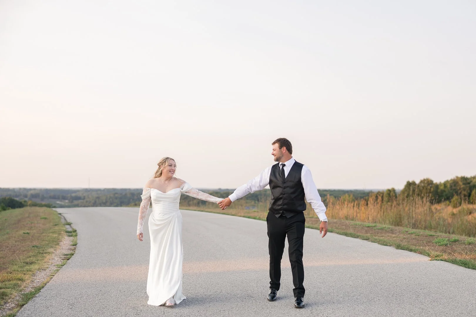 A bride and groom holding hands and walking on a rural road with grass and trees in the background during daytime.