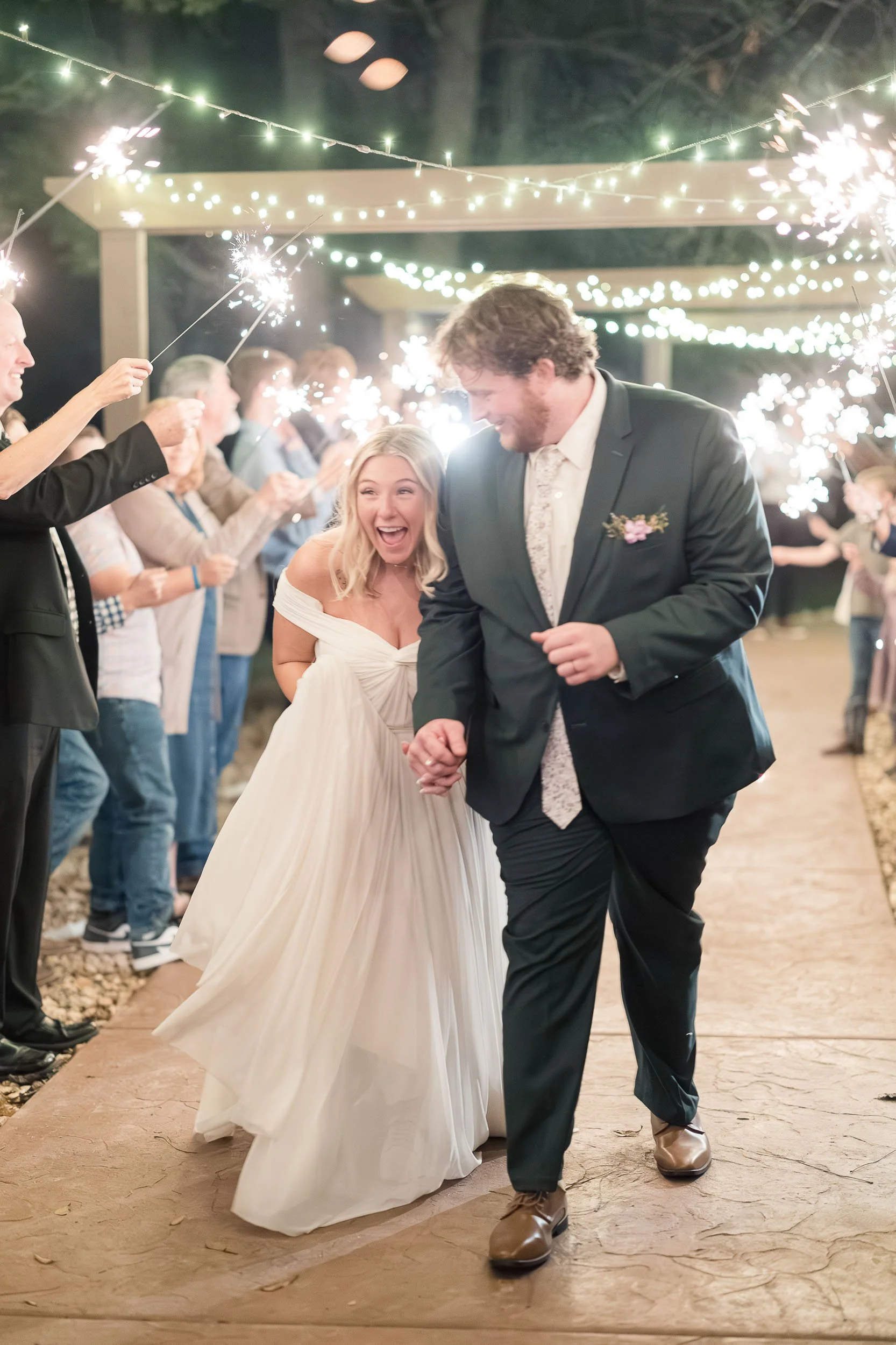 A bride and groom celebrating their wedding night with sparklers while surrounded by guests under string lights outdoors at night.