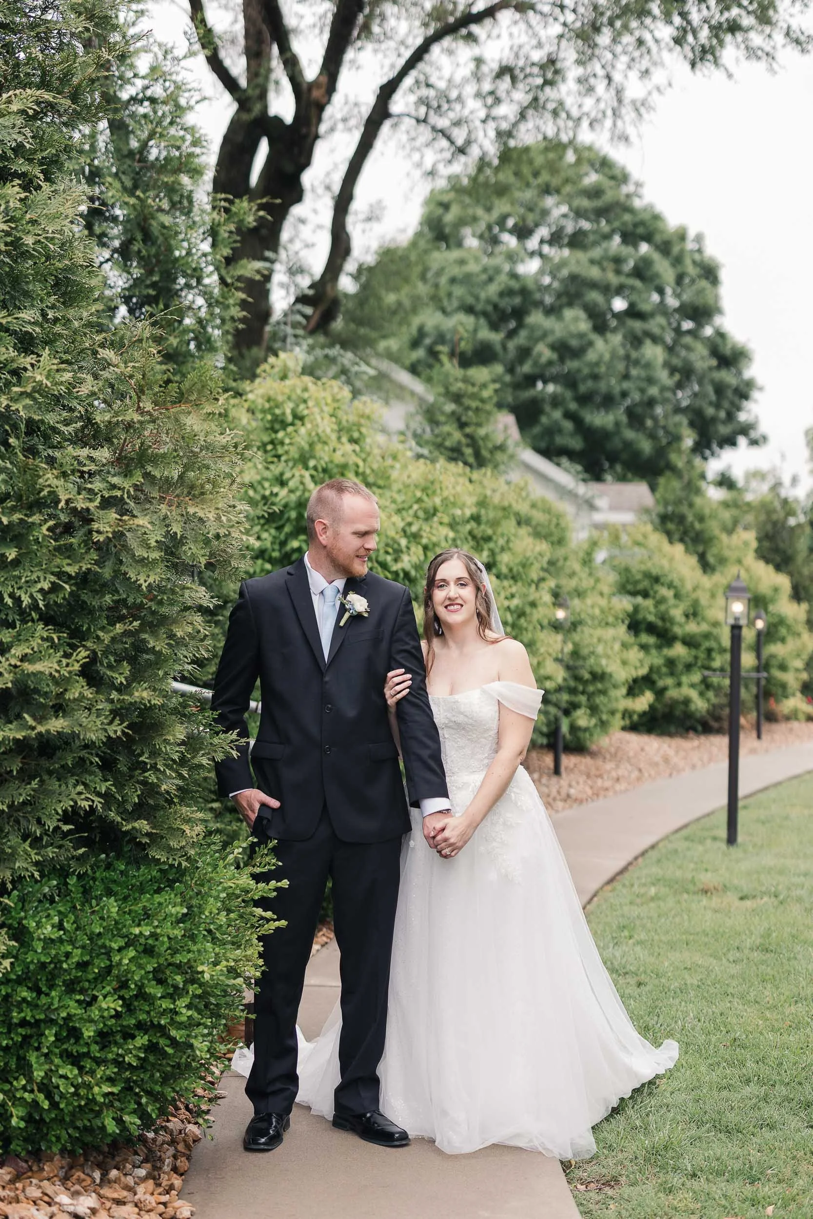 A bride and groom walking arm in arm outdoors on a wedding day, surrounded by greenery, with trees and lamp posts in the background.