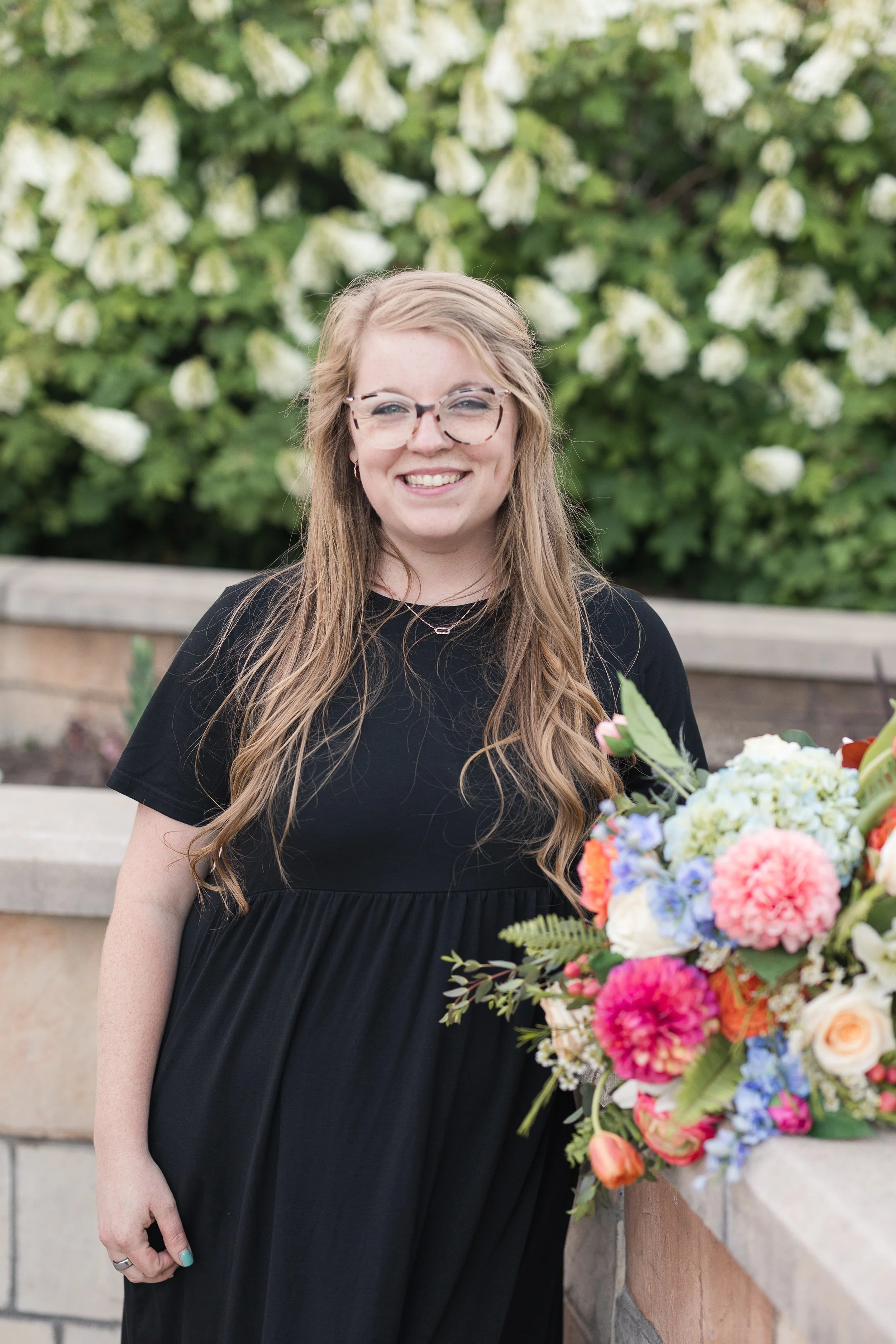 A woman with long wavy blonde hair, wearing glasses and a black dress, smiling and standing outdoors near a colorful flower arrangement, with a background of green foliage and white flowers.