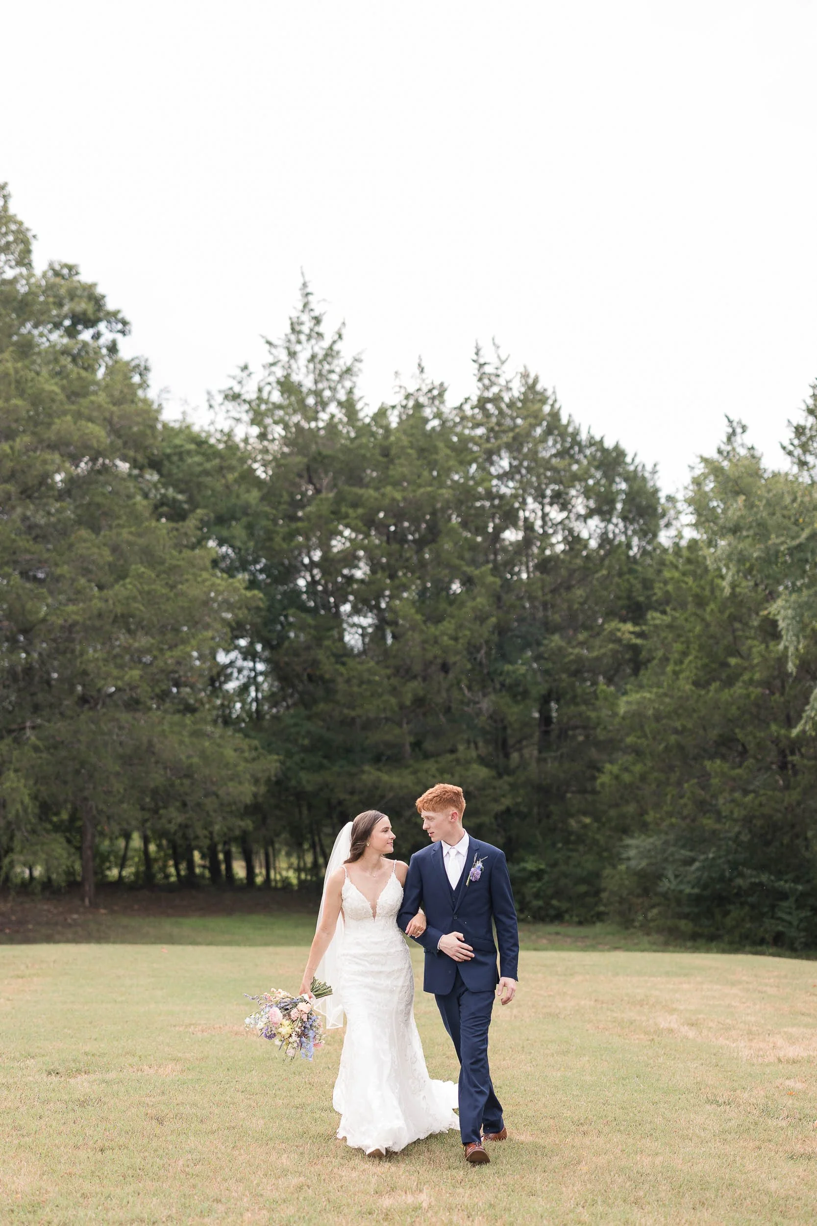 A bride and groom walk arm in arm on a grassy field during their outdoor wedding, surrounded by tall trees and a cloudy sky.