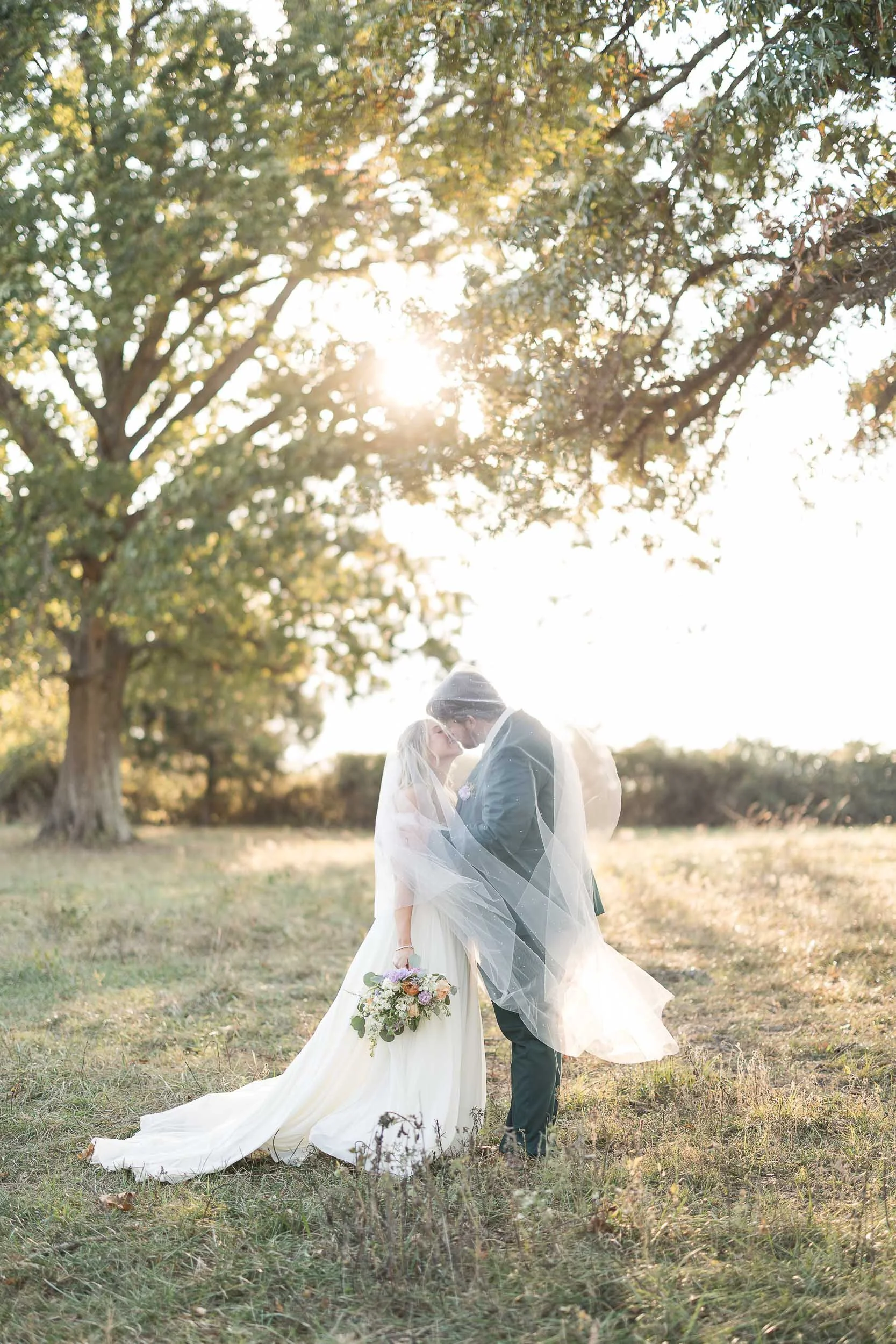 A bride and groom sharing a kiss in a scenic outdoor setting during sunset, with the bride holding a bouquet and wearing a long white gown, and the groom in a dark suit, under a large tree.