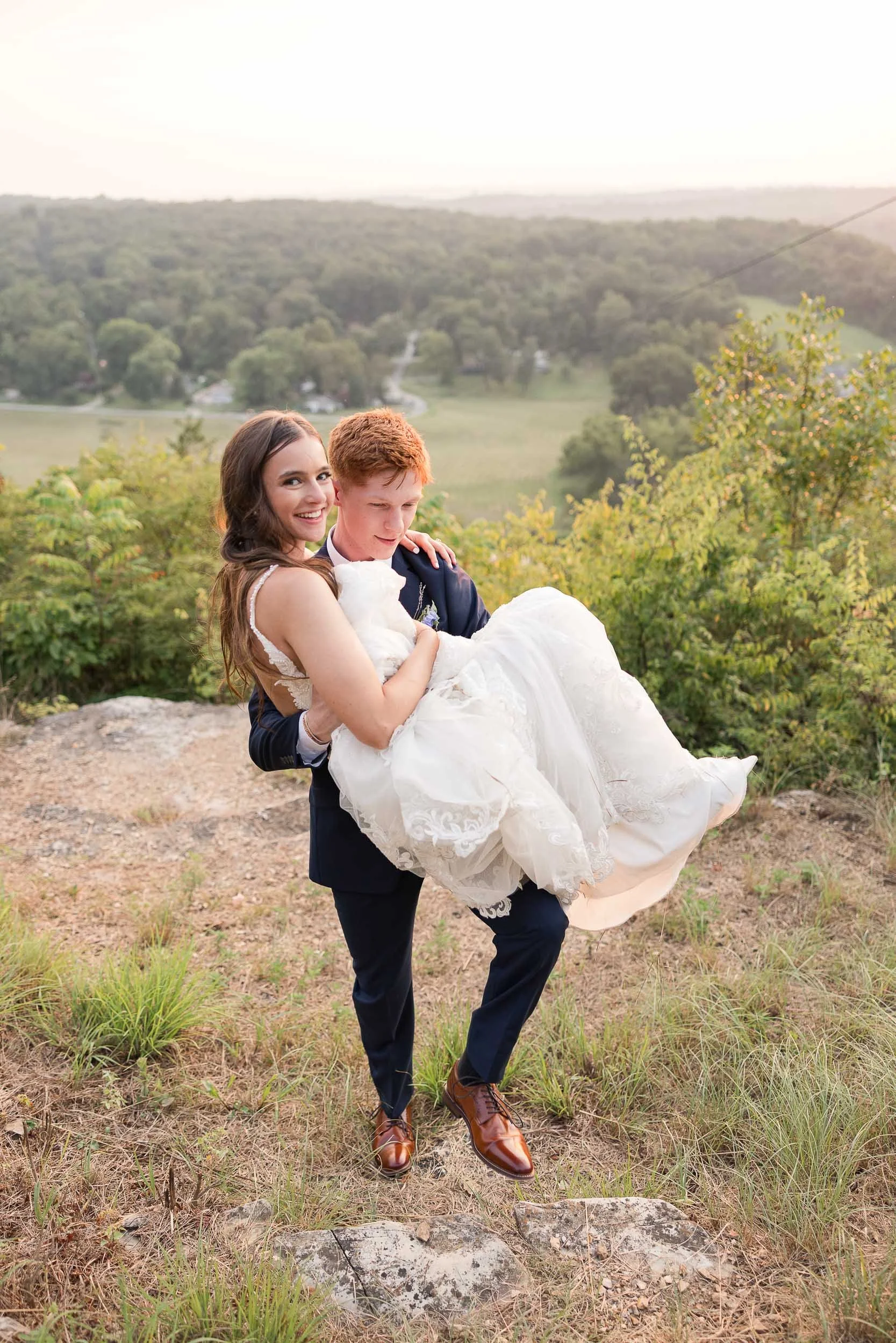 A couple dressed in wedding attire, with the woman in a white wedding dress and the man in a navy suit, standing outdoors in a natural setting with green trees and rolling hills in the background, smiling and holding each other.