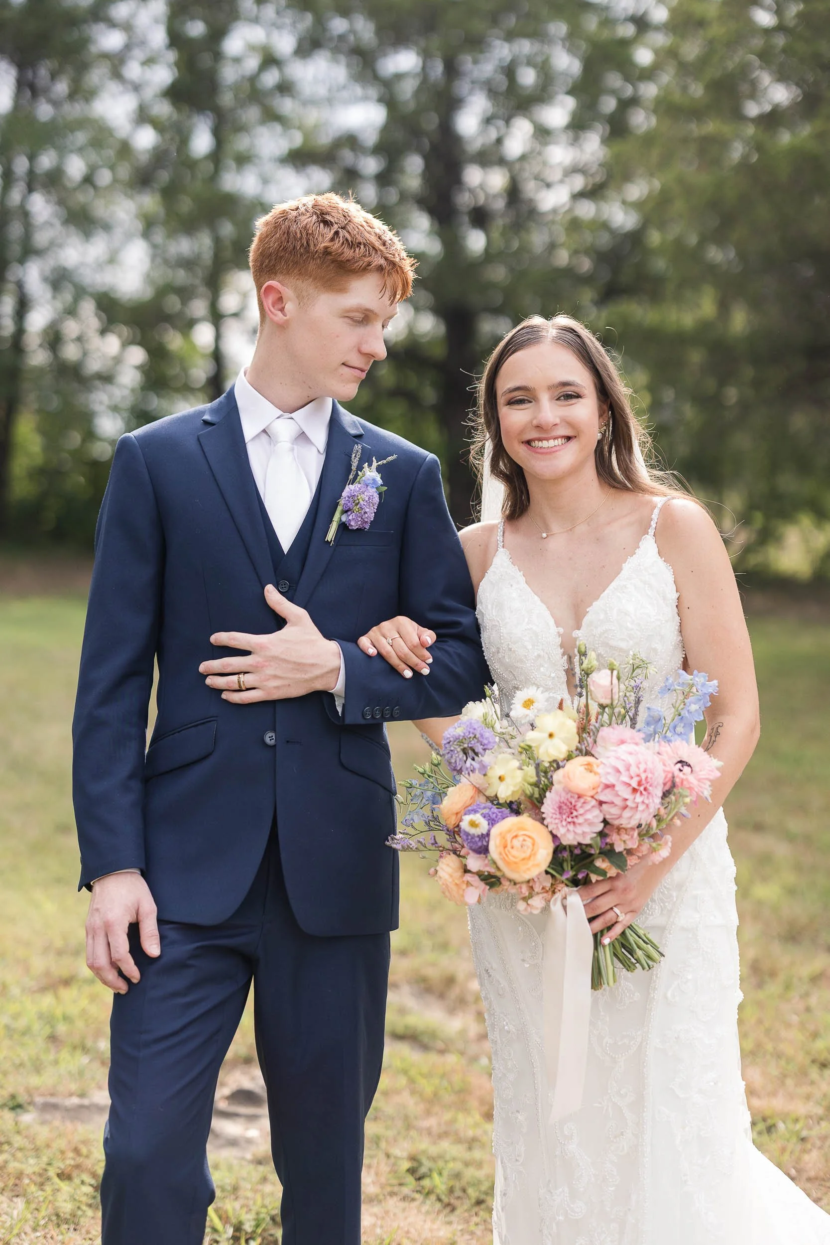 A couple dressed in wedding attire standing outdoors with trees and grass in the background. The man is wearing a navy blue suit and has a purple boutonniere. The woman is wearing a white lace wedding dress and holding a large bouquet of pastel-color