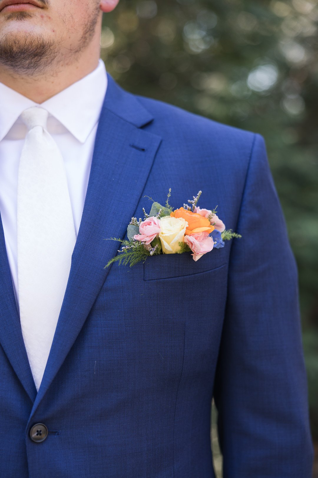 Close-up of a man wearing a navy blue suit jacket with a white shirt and light tie, showing a floral boutonniere on the left lapel.