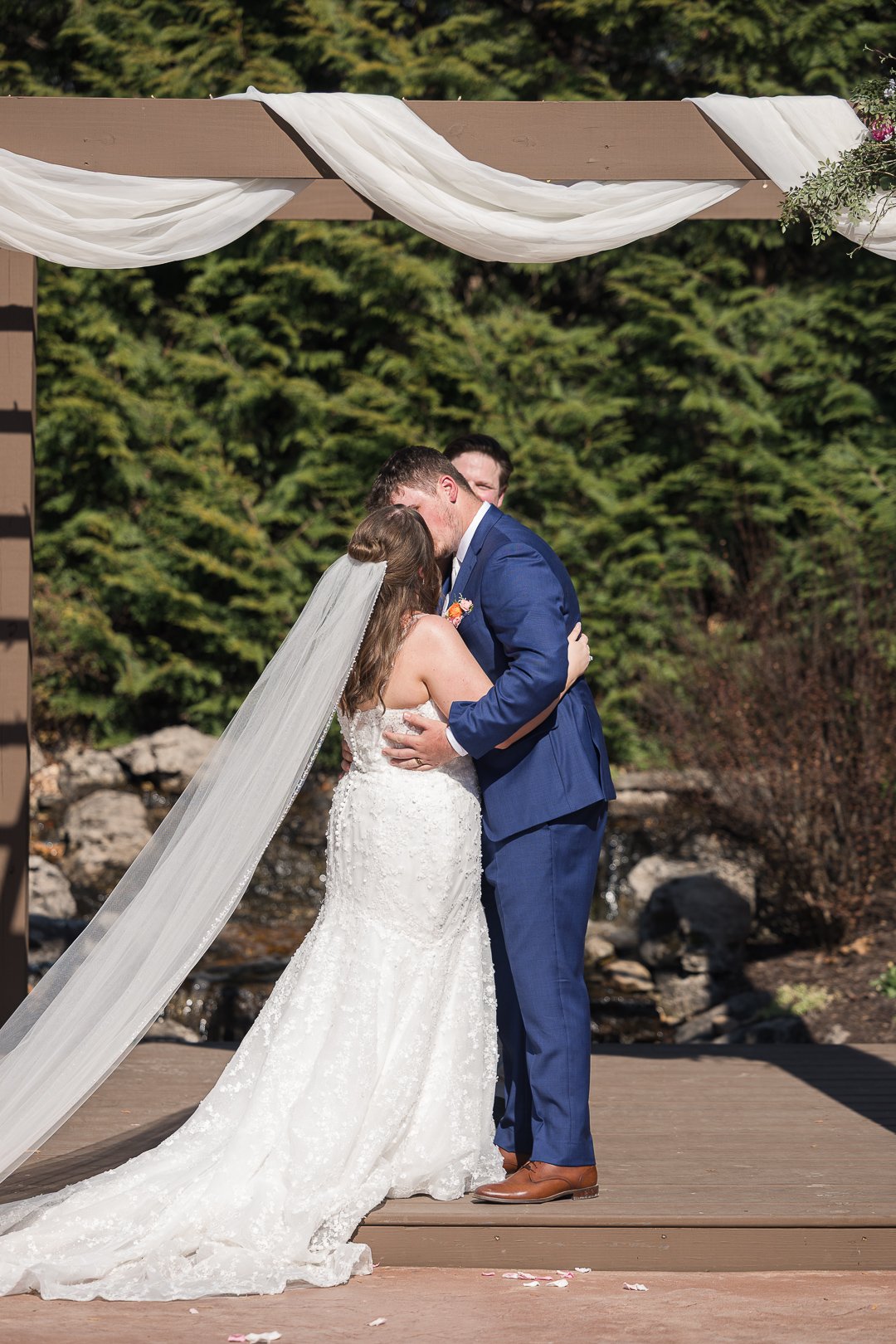 A bride and groom share a kiss during their outdoor wedding ceremony, standing on a wooden platform with greenery and rocks in the background.