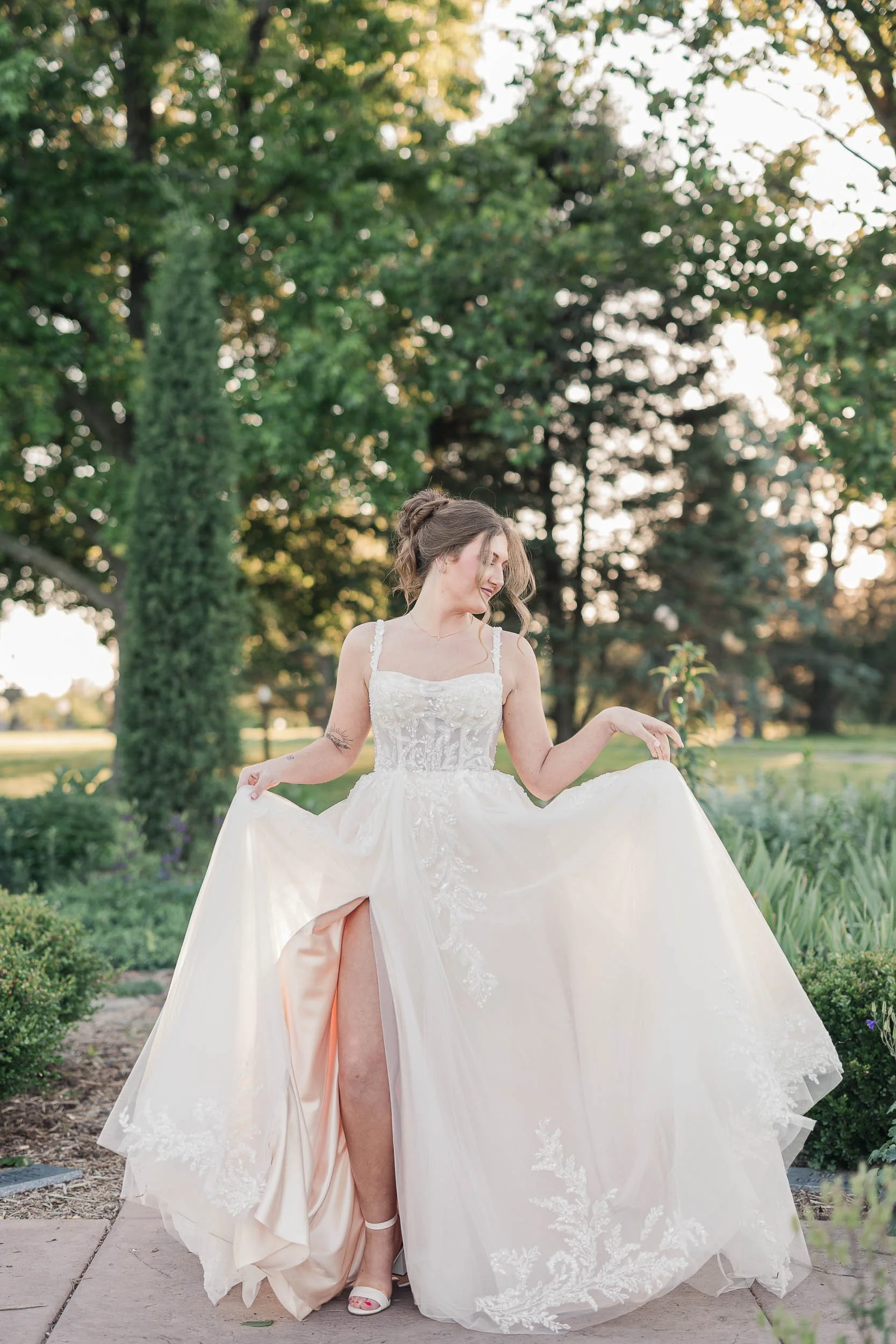 A woman in a wedding dress walking outdoors in a park with trees in the background, lifting her dress to show her shoes and a tattoo on her arm.