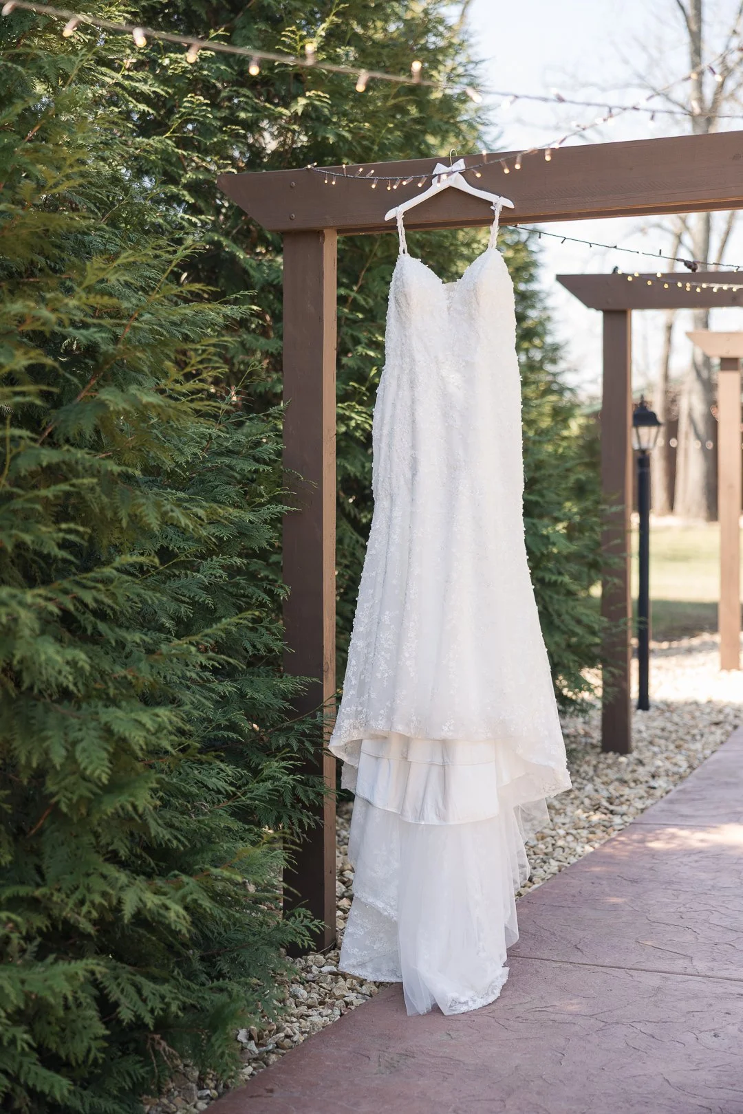 White wedding dress hanging on a hanger outdoors on a wooden structure with string lights, surrounded by green foliage and trees.