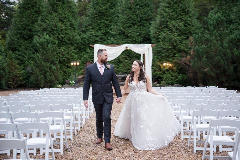 Outdoor wedding ceremony setup with bride and groom walking down aisle, surrounded by white chairs and greenery backdrop.