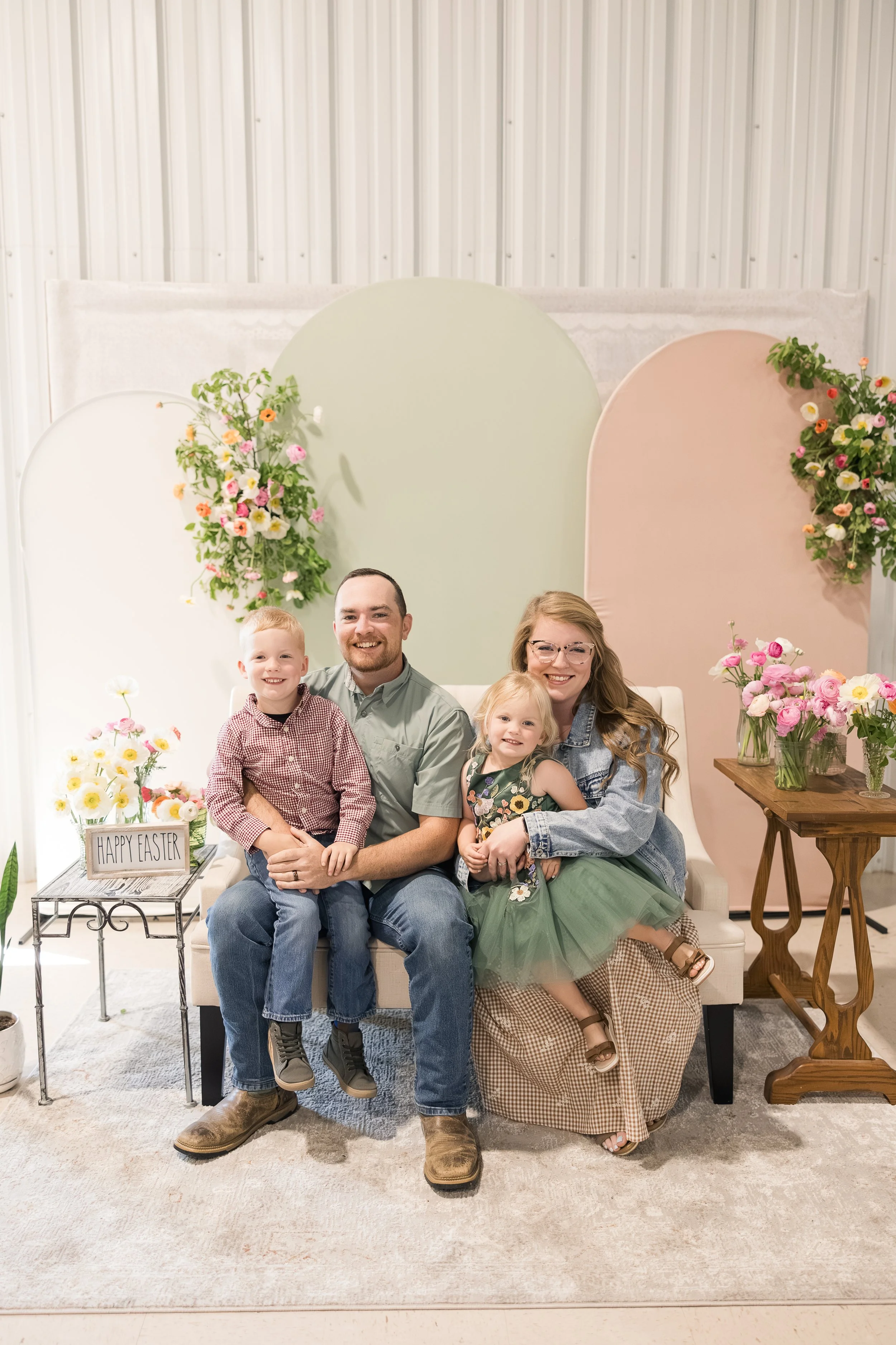 Family sitting on a beige couch with a floral backdrop. A man and a woman sit with three children, smiling. The setting features floral arrangements and Easter decorations, including a sign that reads 'Happy Easter'.