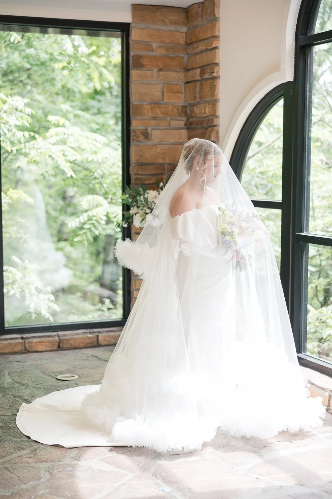 A bride in a white wedding gown and veil standing indoors, holding a bouquet, looking out a large window with greenery outside, with a brick wall and stone floor in the background.
