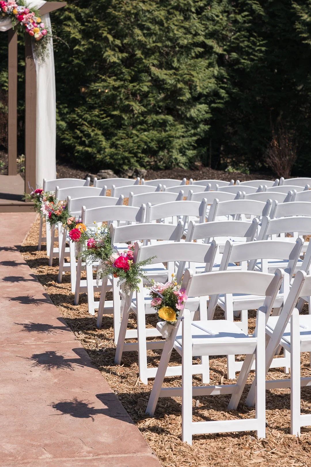 Wedding ceremony setup with white chairs decorated with colorful flowers, located outdoors on a sunny day with green trees in the background.