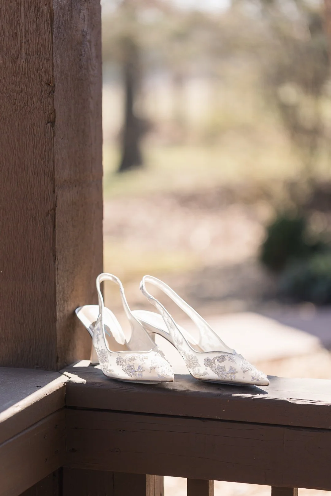 A pair of white lace wedding shoes with pointed toes and slingback straps, placed on a wooden ledge outside with blurred trees in the background.