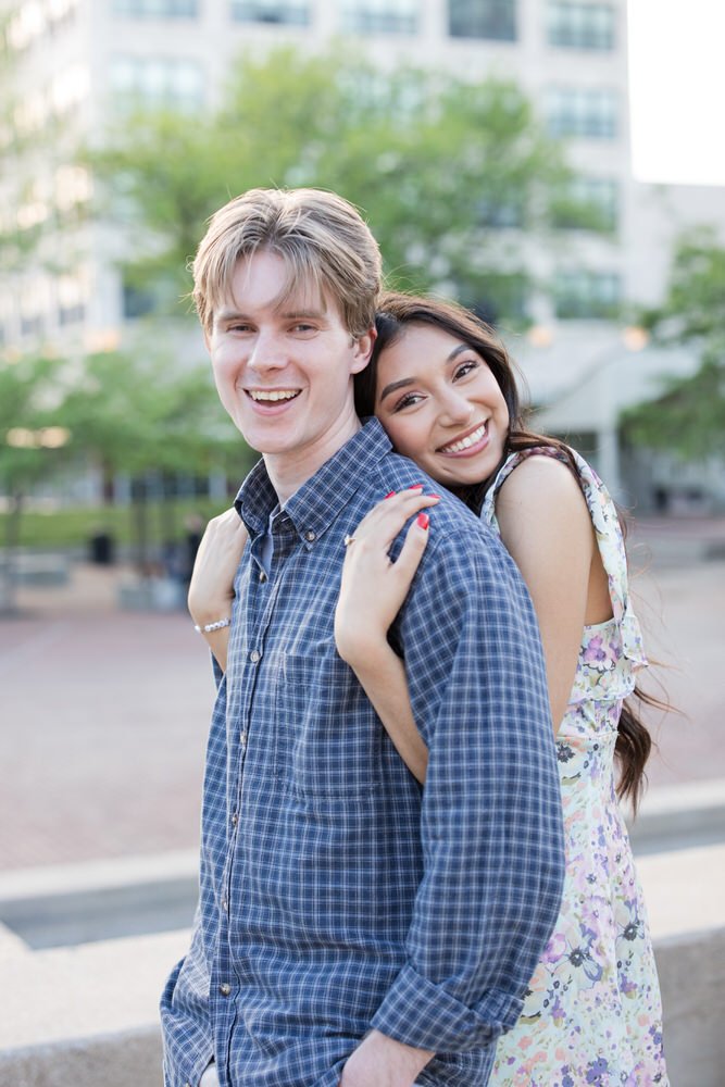 Smiling couple posing outdoors with trees and a building in the background.