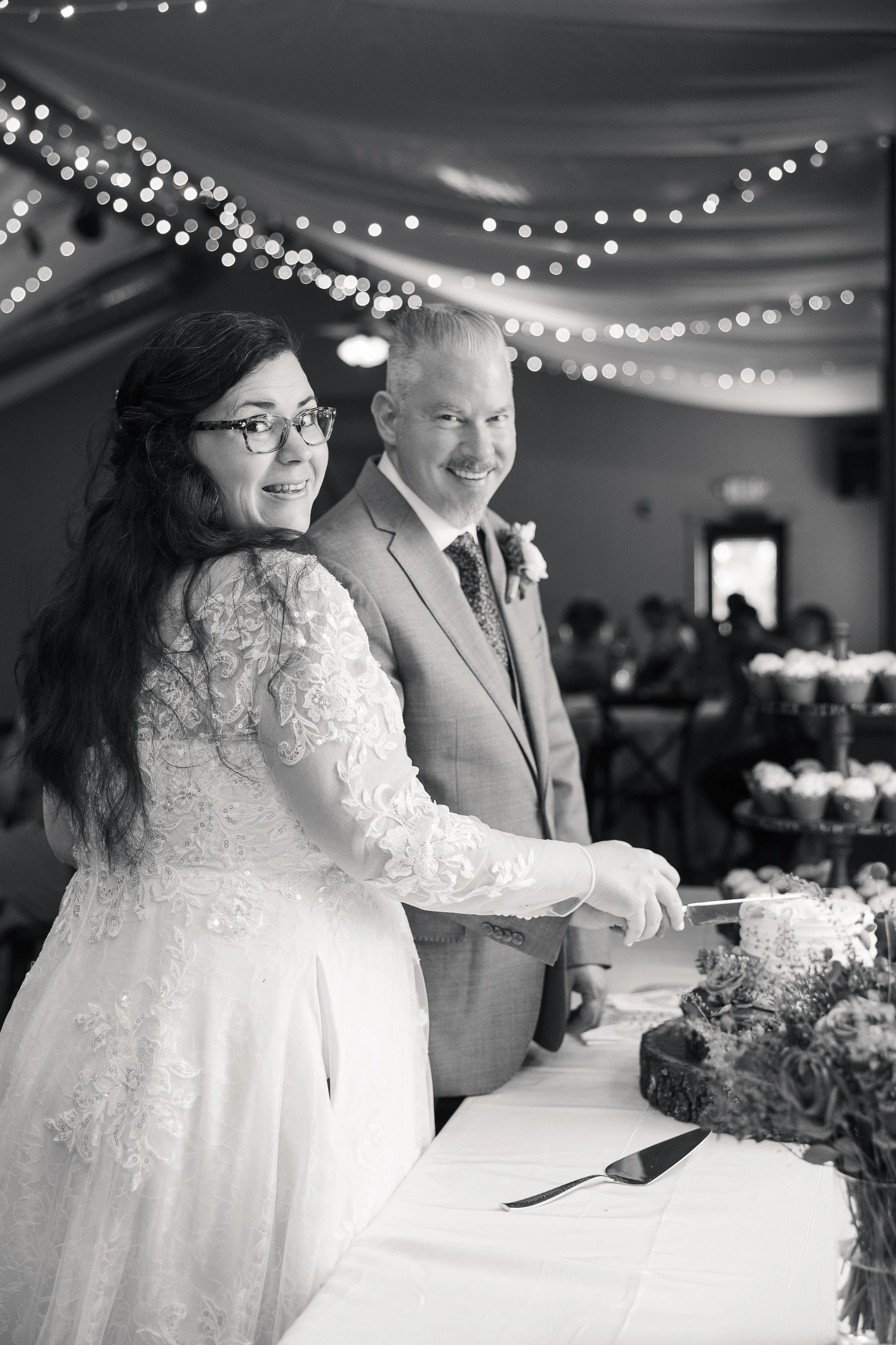 A black-and-white photo of a bride and groom smiling while cutting their wedding cake, decorated with flowers, under string lights in a decorated reception hall.