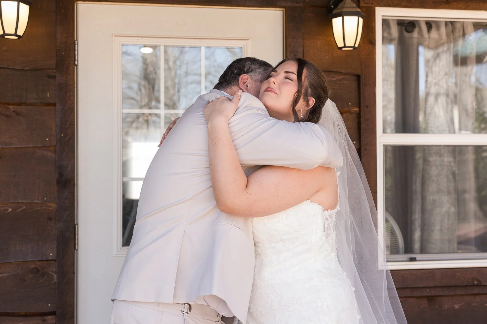 A groom hugging a bride in a white wedding dress and veil inside a room with wooden walls and windows, during daytime.