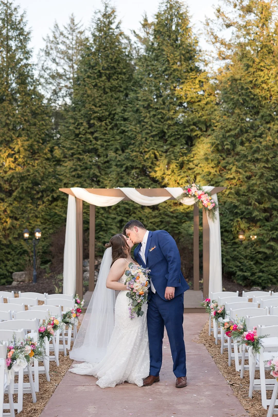 A bride and groom share a kiss during their outdoor wedding ceremony. The bride wears a white wedding gown and holds a colorful bouquet, and the groom wears a blue suit. They stand under a wooden wedding arch draped with white fabric and pink flowers