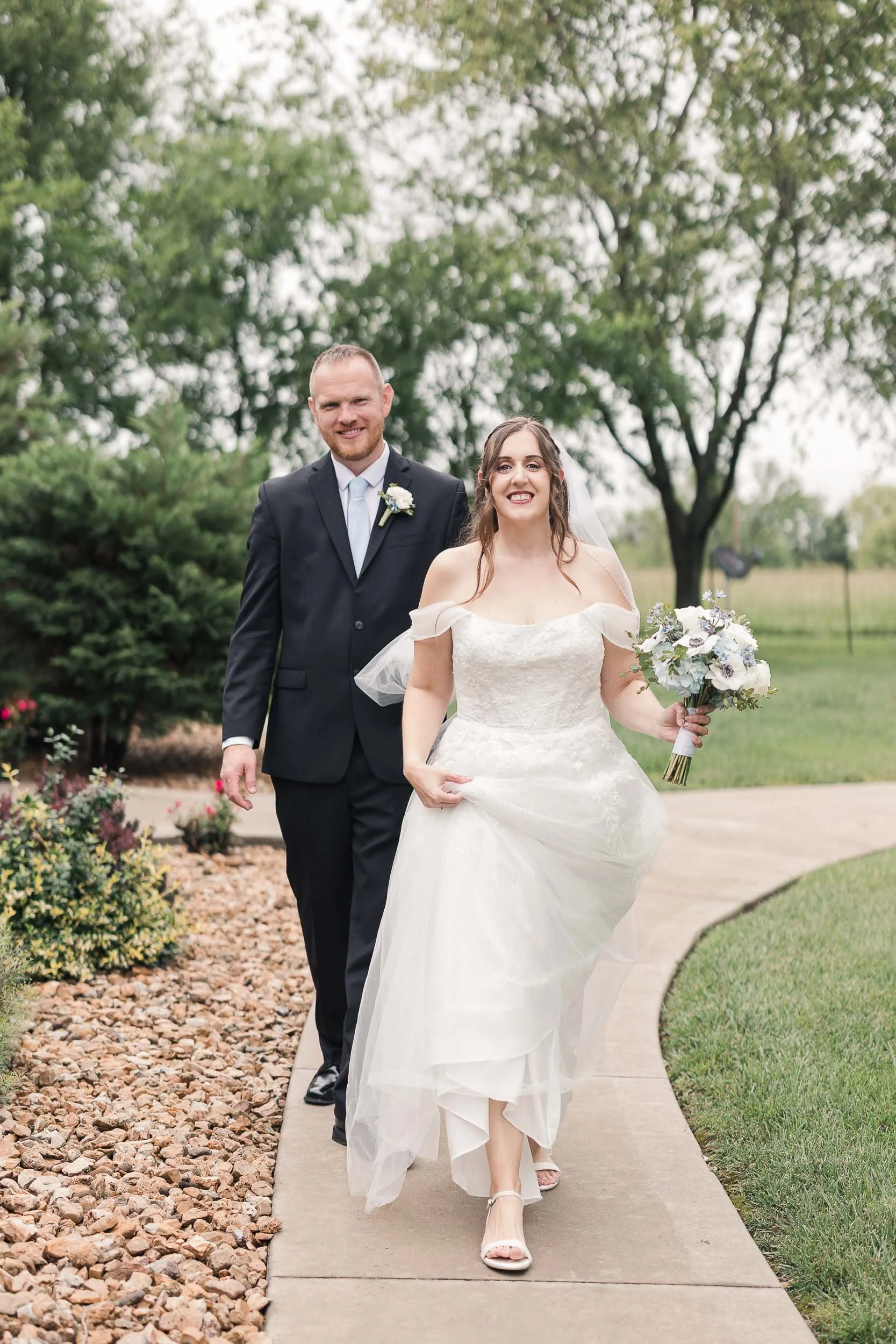 A newlywed couple walking outdoors on a paved path, with the bride holding a bouquet of flowers, and the groom dressed in a black suit and tie, smiling at the camera, in a garden setting with trees and bushes in the background.