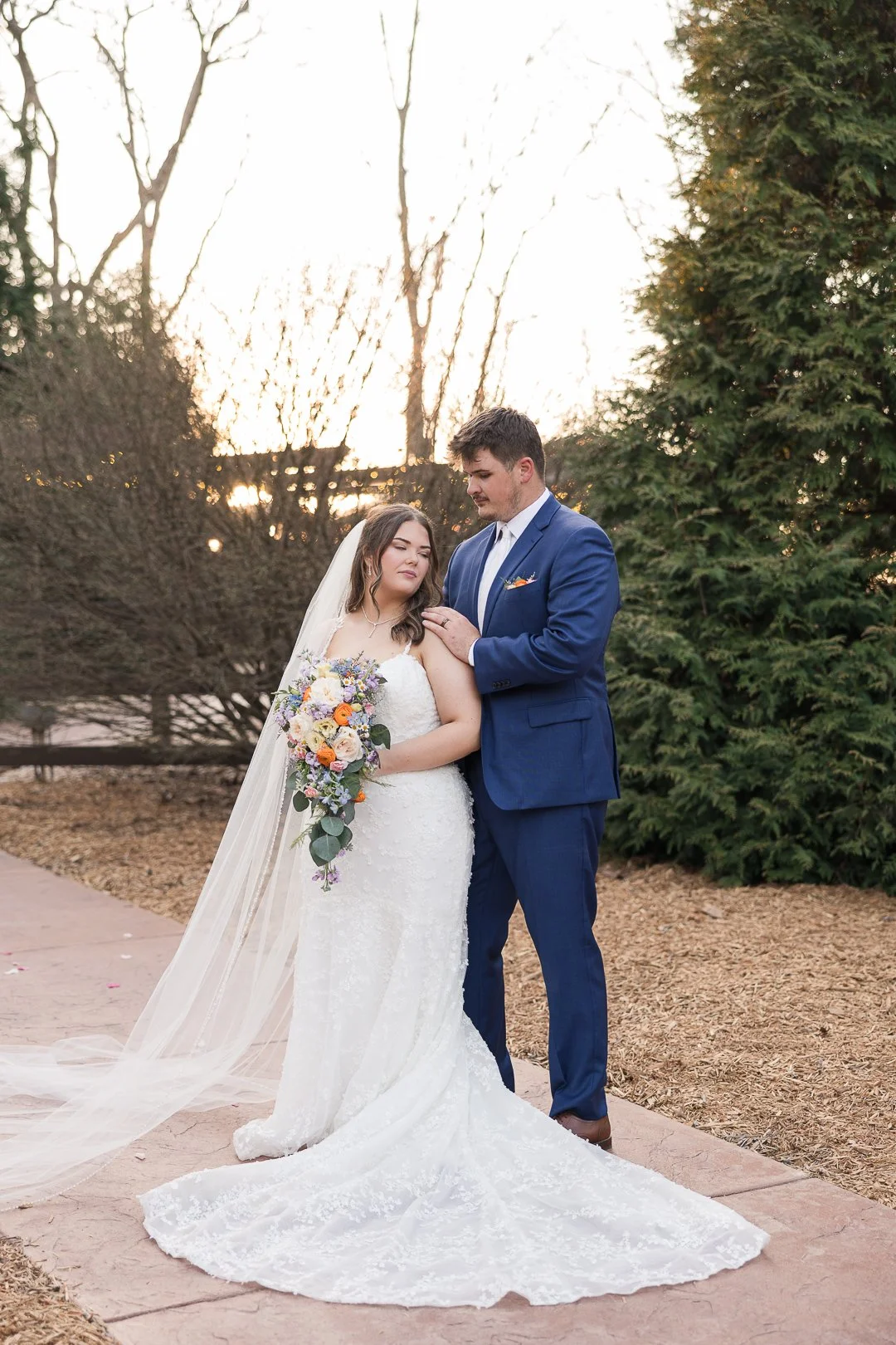 A bride and groom standing outdoors in a garden during sunset, with the bride holding a bouquet of flowers and the groom gently touching her shoulder.