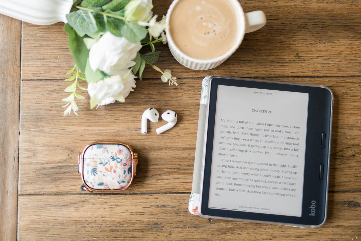 A flat lay of a wooden table with a plant, a mug of coffee, wireless earbuds, a floral case, and an e-reader displaying a book.