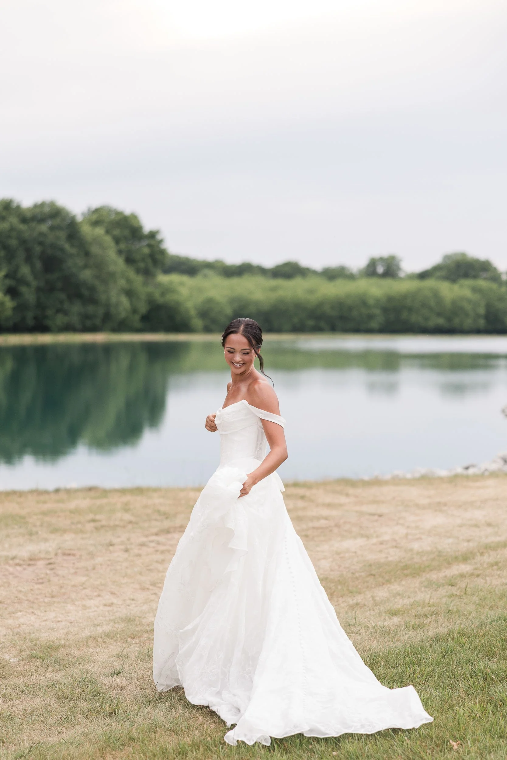 A smiling woman in a white wedding dress standing on grass near a body of water with trees in the background during daytime.
