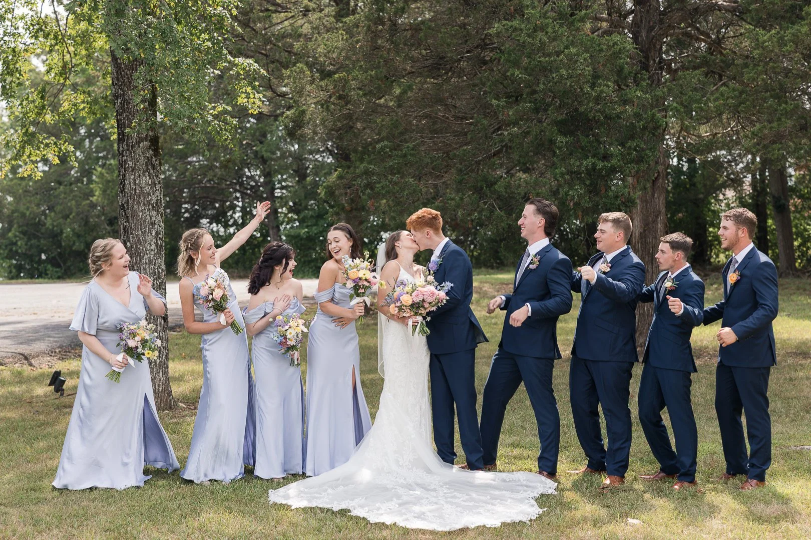 A wedding party outdoors with a bride and groom sharing a kiss, surrounded by bridesmaids and groomsmen celebrating and smiling, with trees in the background.