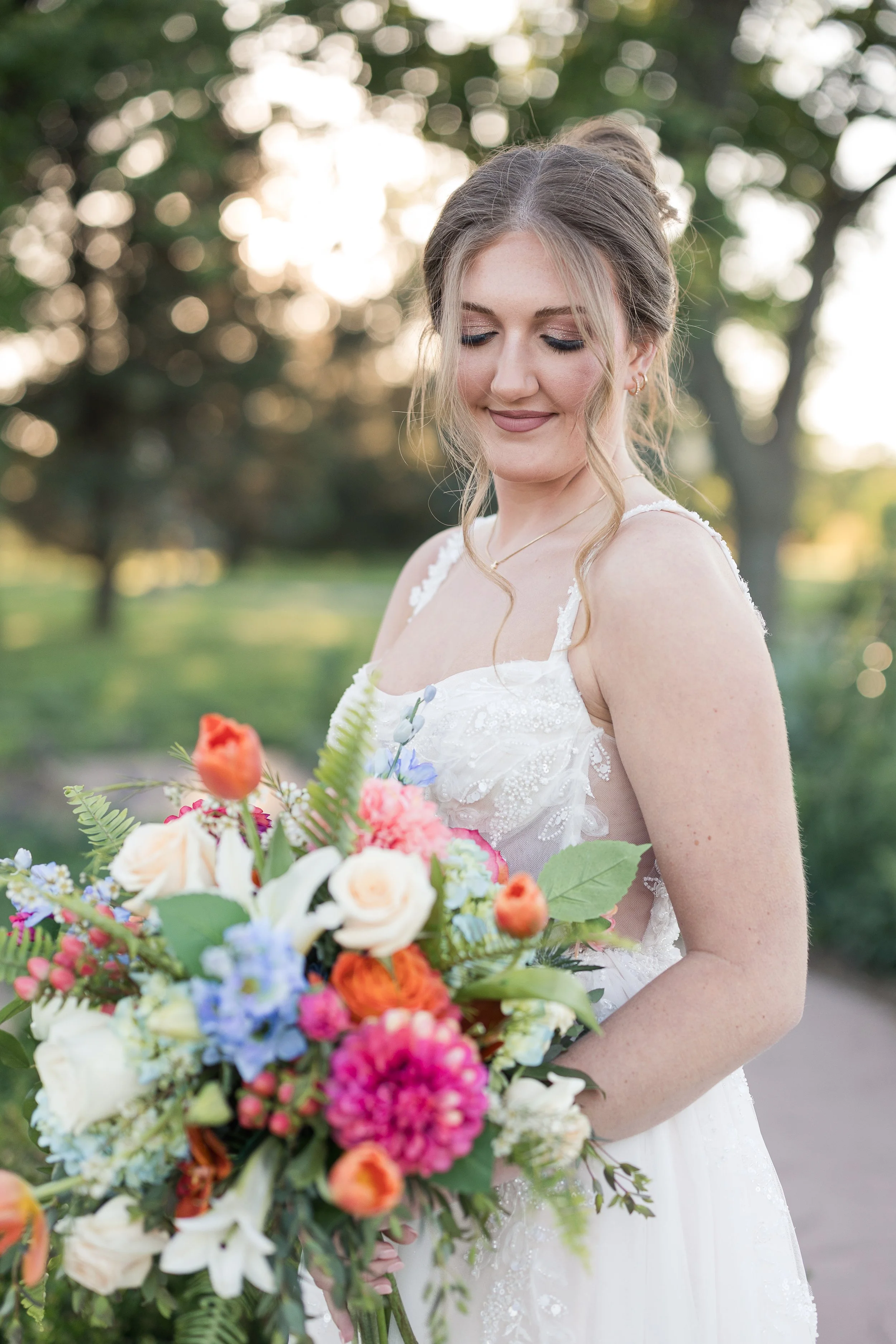A bride in a white wedding dress holding a colorful bouquet of flowers outdoors during sunset, with trees and soft sunlight in the background.