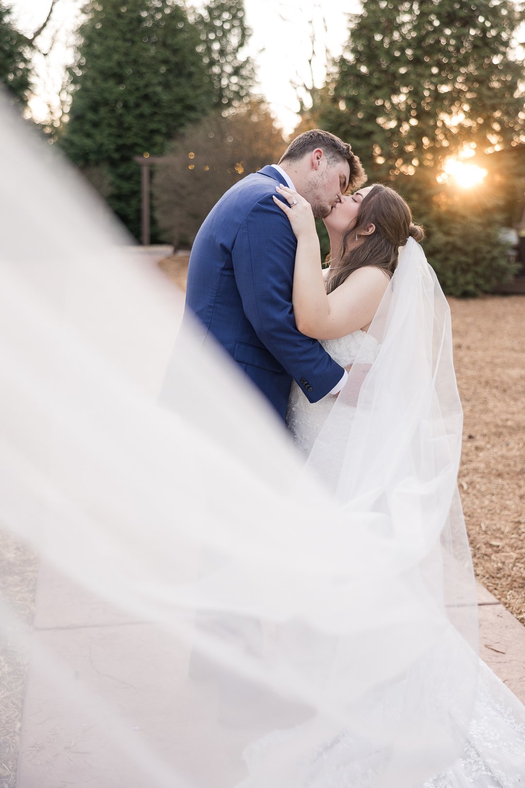 A bride and groom sharing a kiss outdoors at sunset, with trees in the background and a white veil flowing in the foreground.