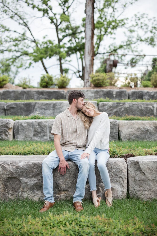 Couple sitting on stone steps in park, man kissing woman's forehead.
