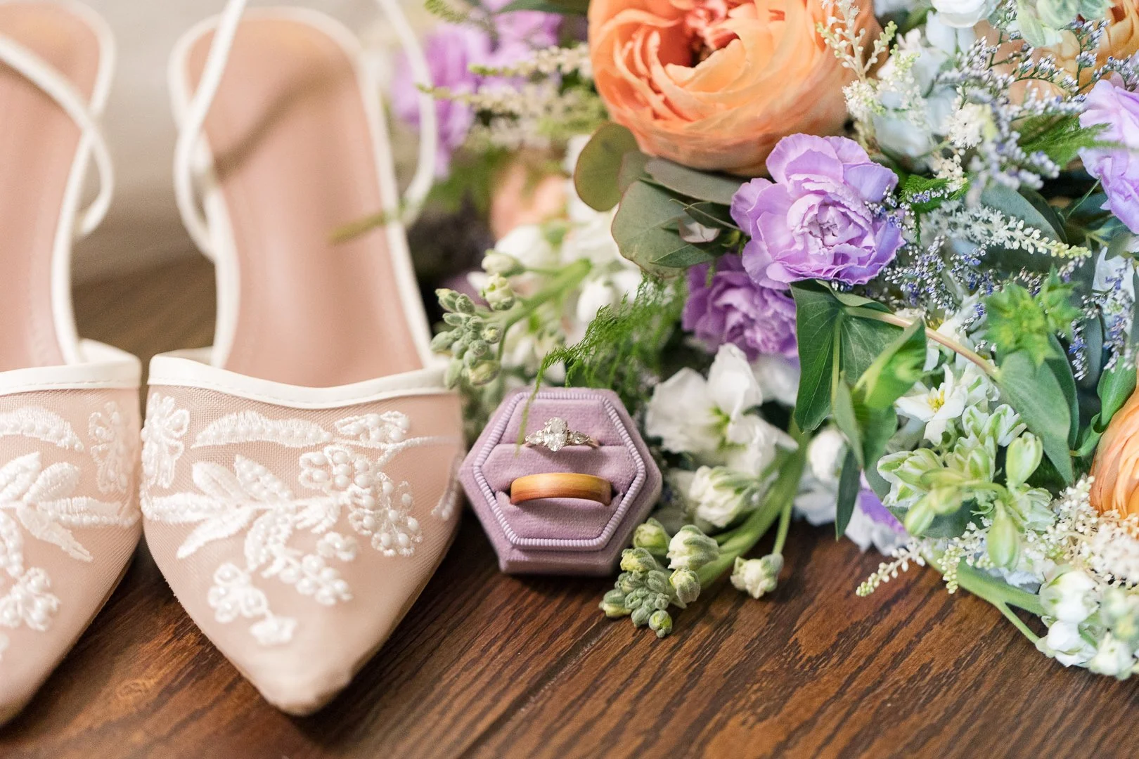A pair of pink embroidered shoes, a lavender ring box with engagement rings, and a bouquet of pink, purple, and white flowers on a wooden surface.