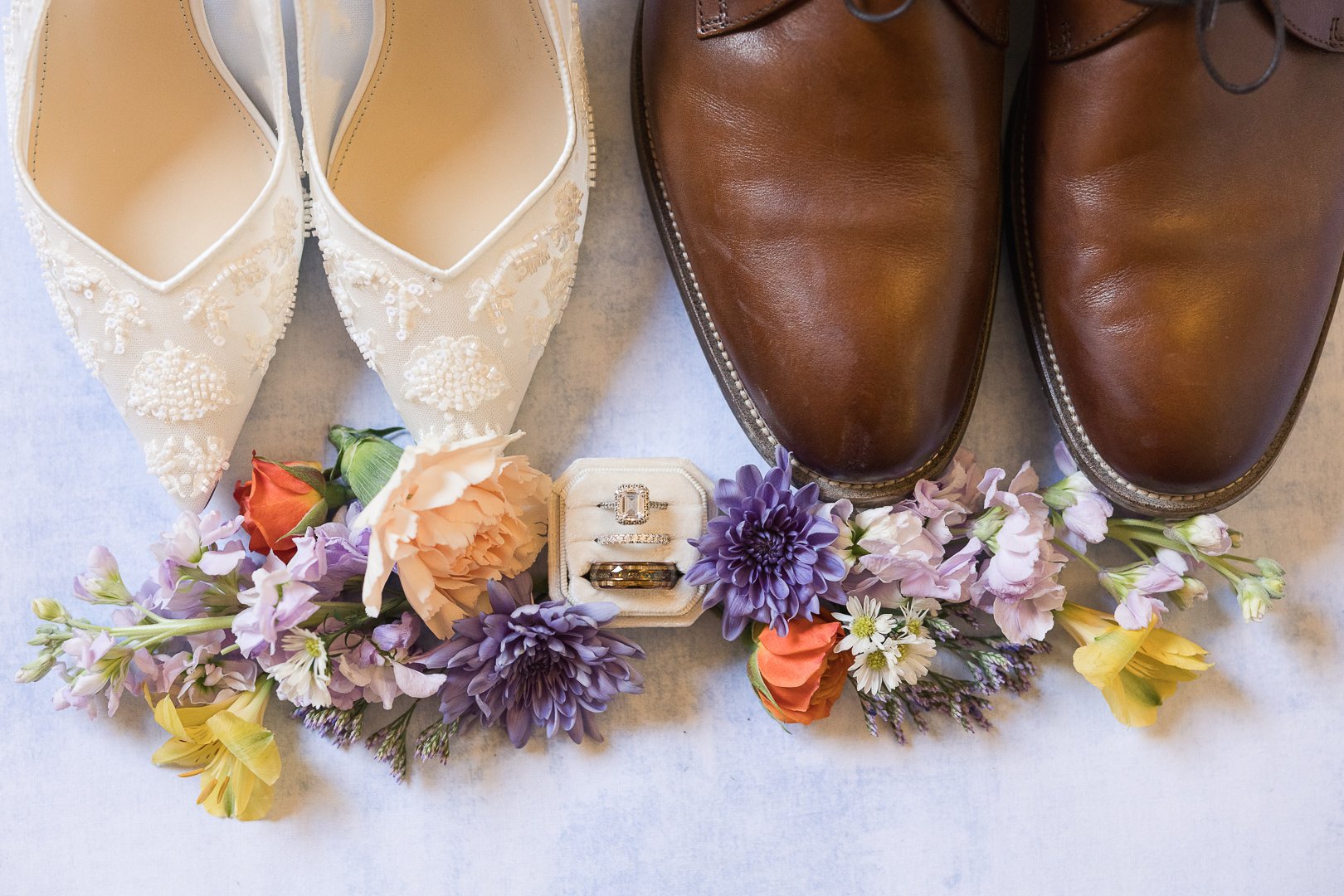 A flat lay of wedding accessories including white embroidered shoes, brown leather shoes, a box of rings, and colorful flowers.