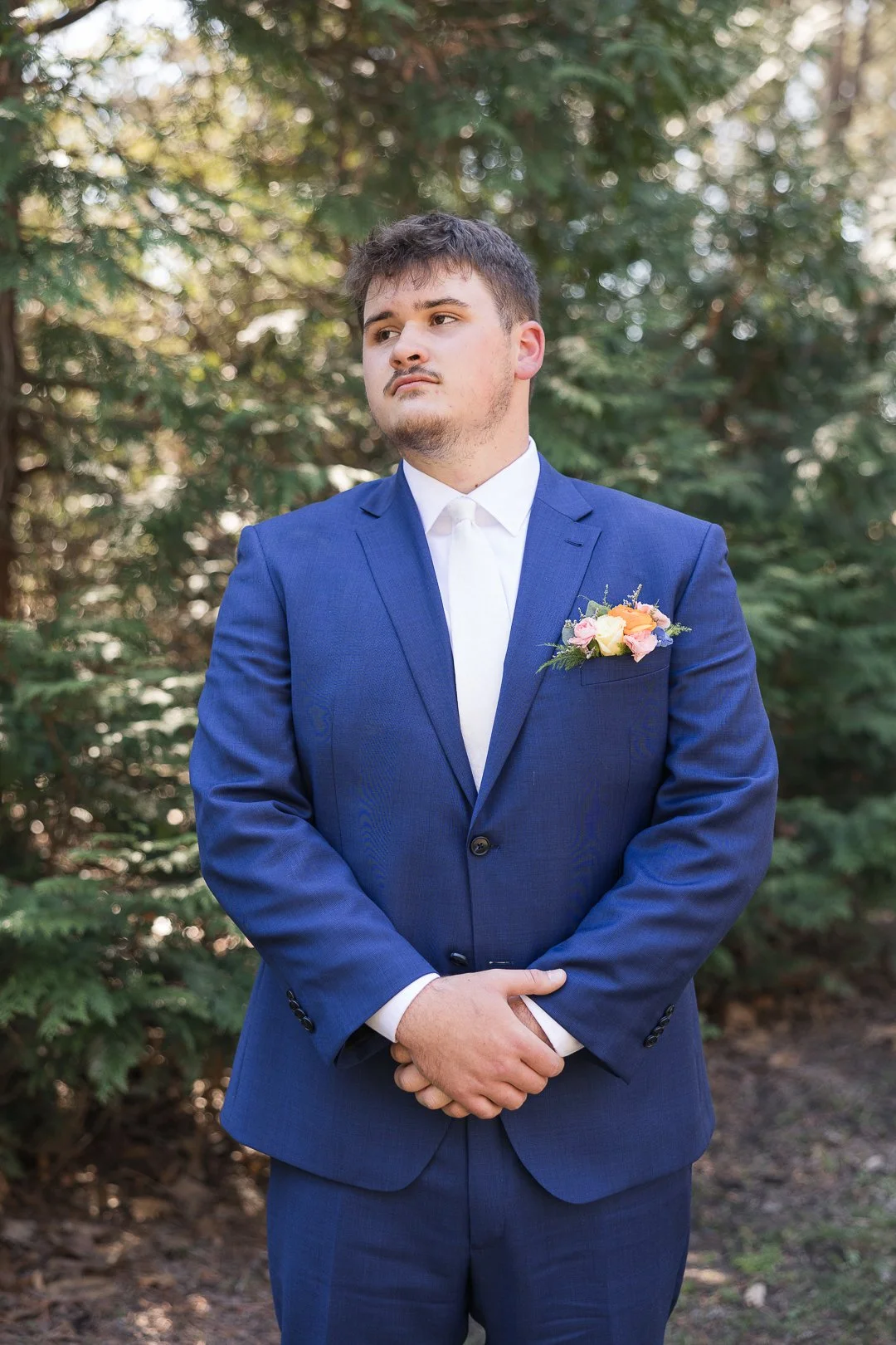 A young man in a blue suit with a white shirt and a boutonniere on his lapel stands outdoors with trees in the background, looking to his left with a serious expression.