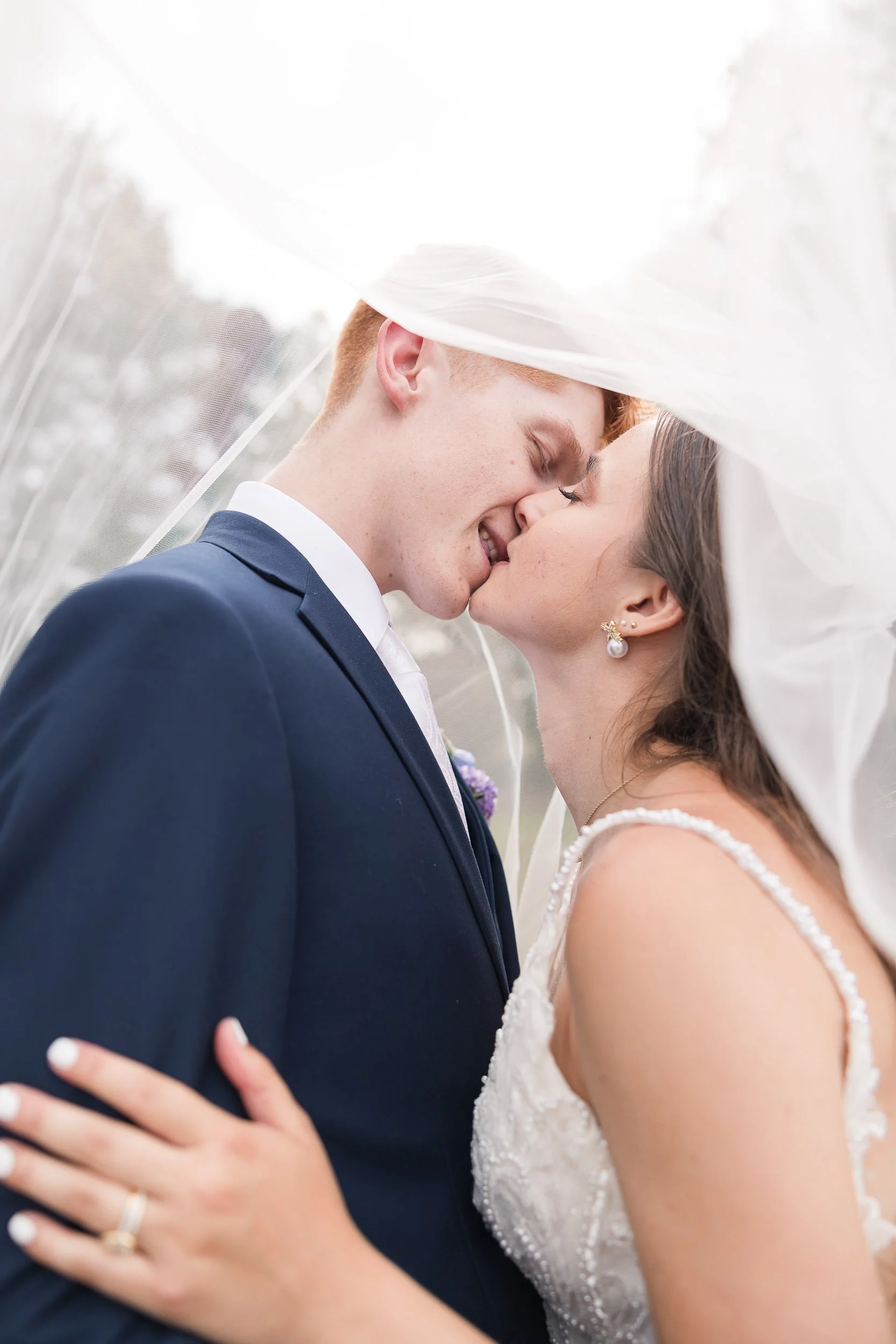 A bride and groom sharing a kiss under a sheer white veil, with close-up focus on their faces and hands, on their wedding day.