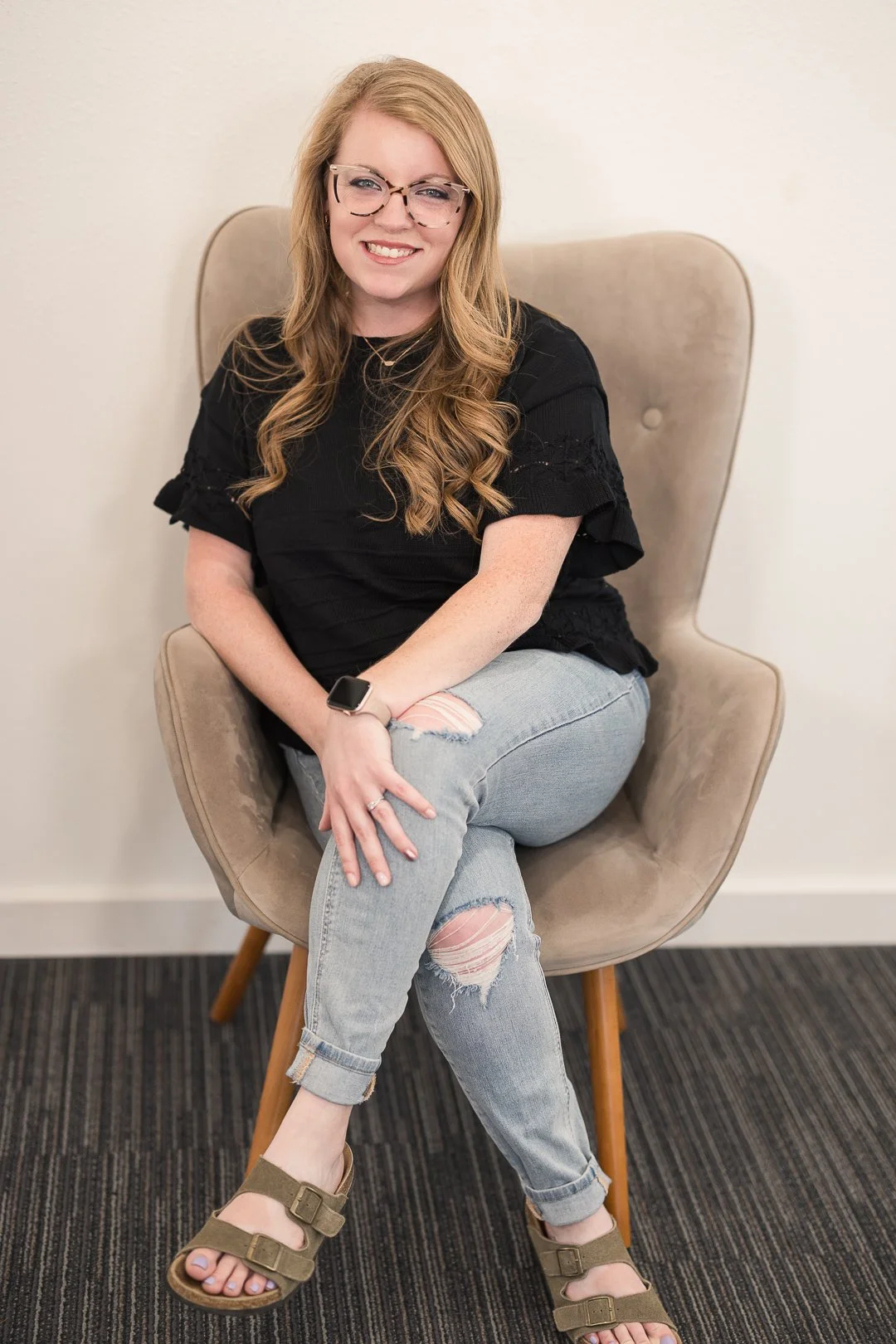 A young woman with long wavy blonde hair, wearing glasses, a black top, ripped jeans, and sandals, sitting on a beige chair against a plain white wall.
