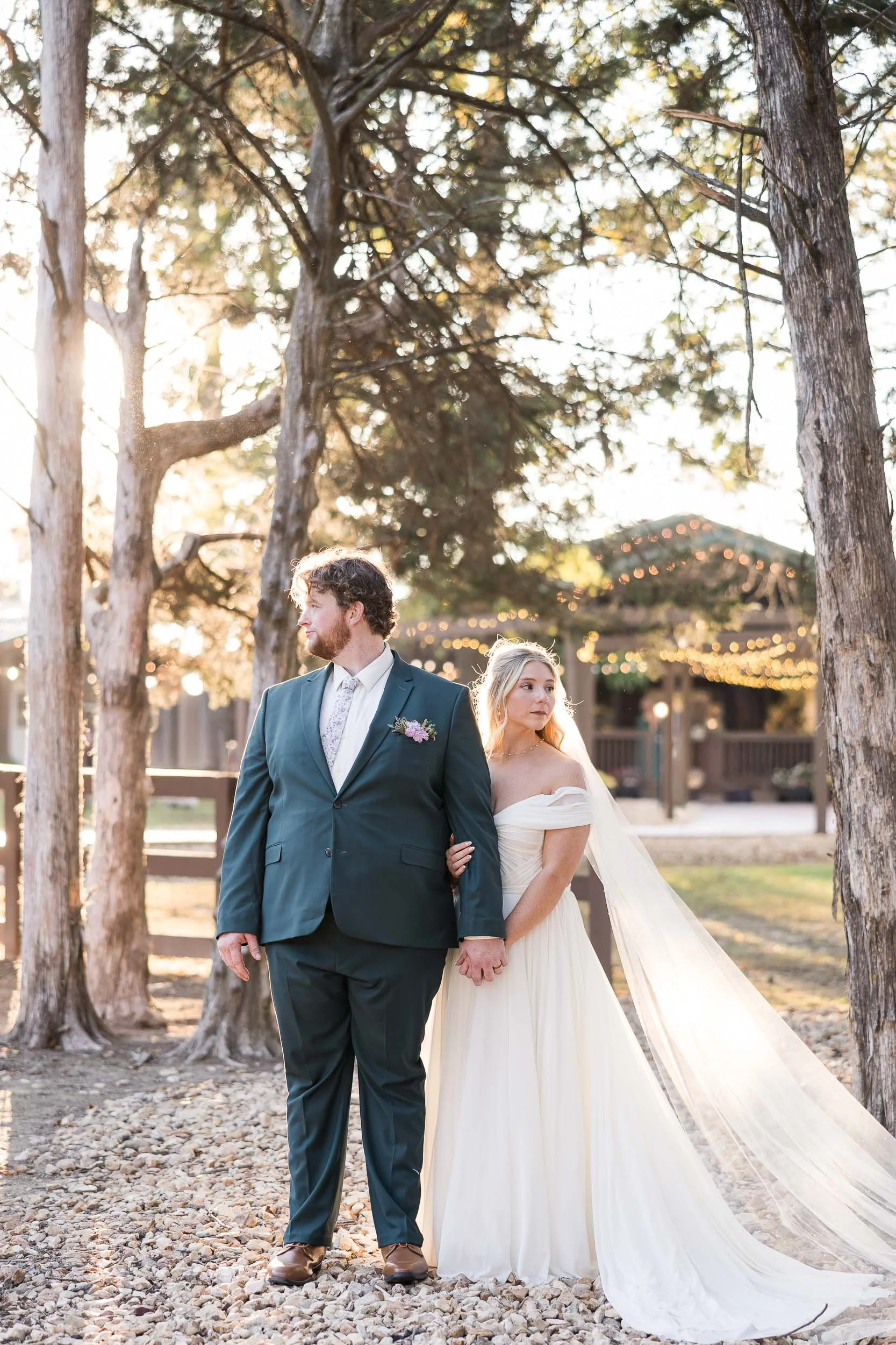 A bride and groom holding hands, standing outdoors among trees with sunlight filtering through, during a wedding ceremony.