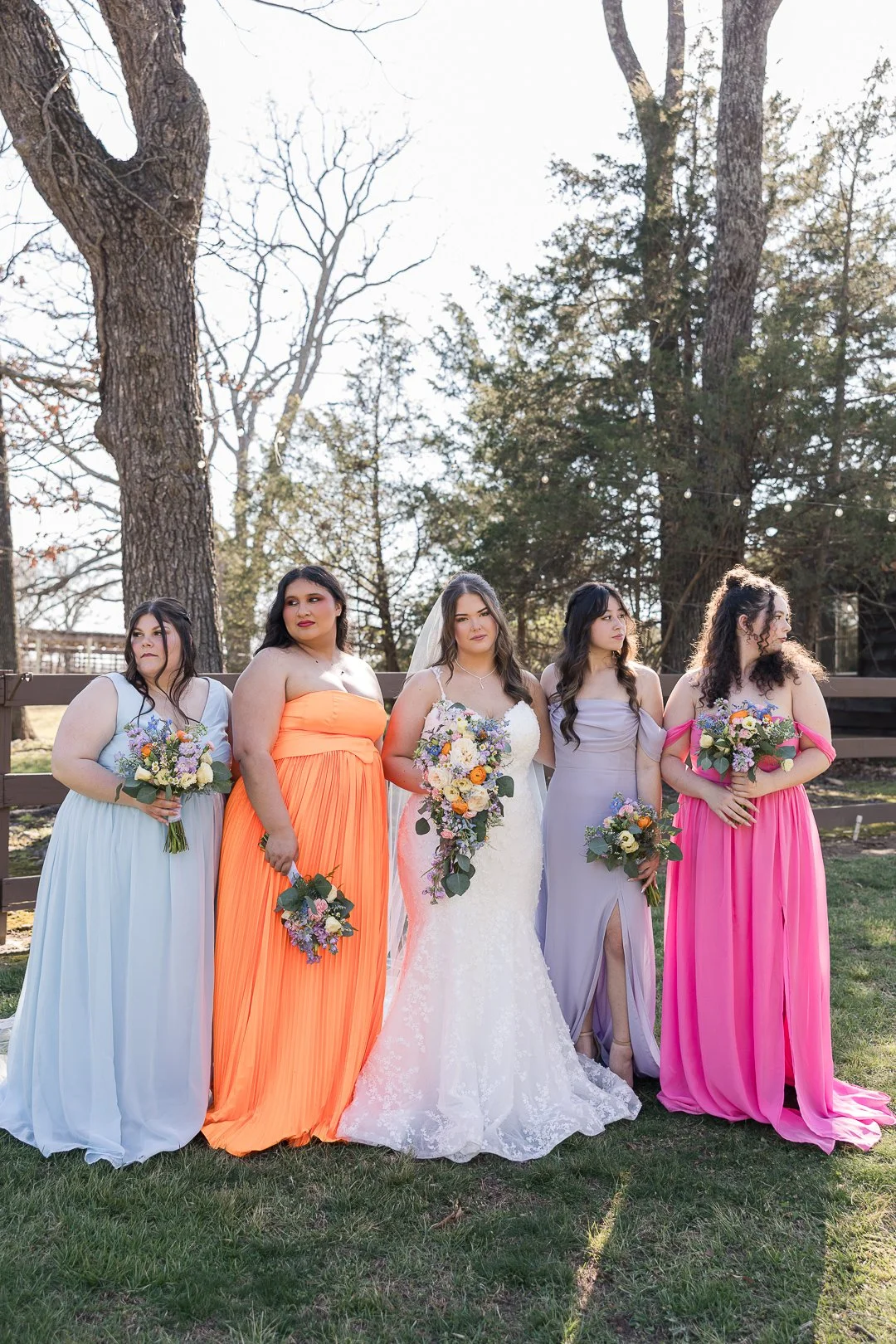 A bride and four bridesmaids standing outdoors in a line during daytime, with trees and string lights in the background. The bride wears a white lace wedding dress with a veil, and the bridesmaids wear colorful dresses in pastel blue, bright orange, 