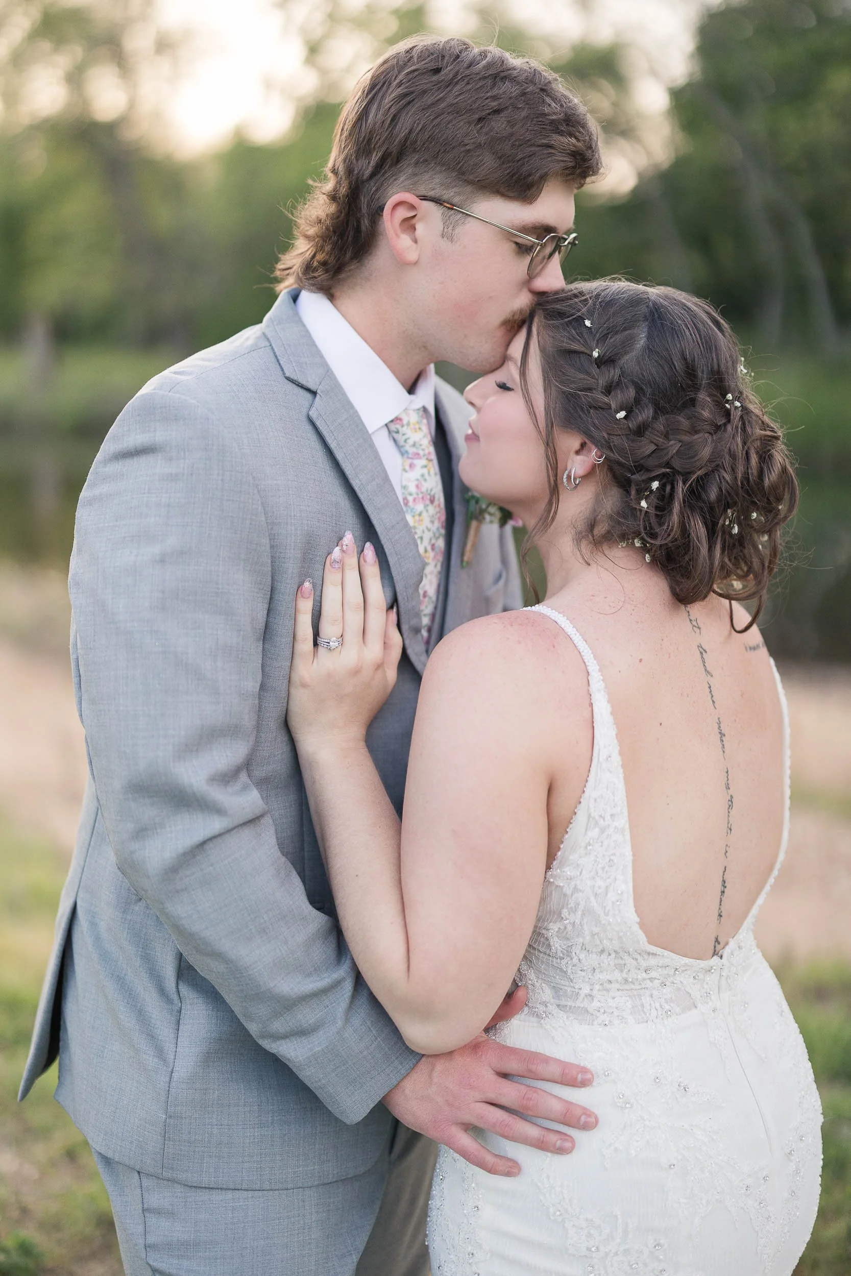 A bride and groom share a kiss in a forest setting, with the bride holding a bouquet of white flowers, and the bride wearing a white wedding gown and veil, and the groom dressed in a beige suit.