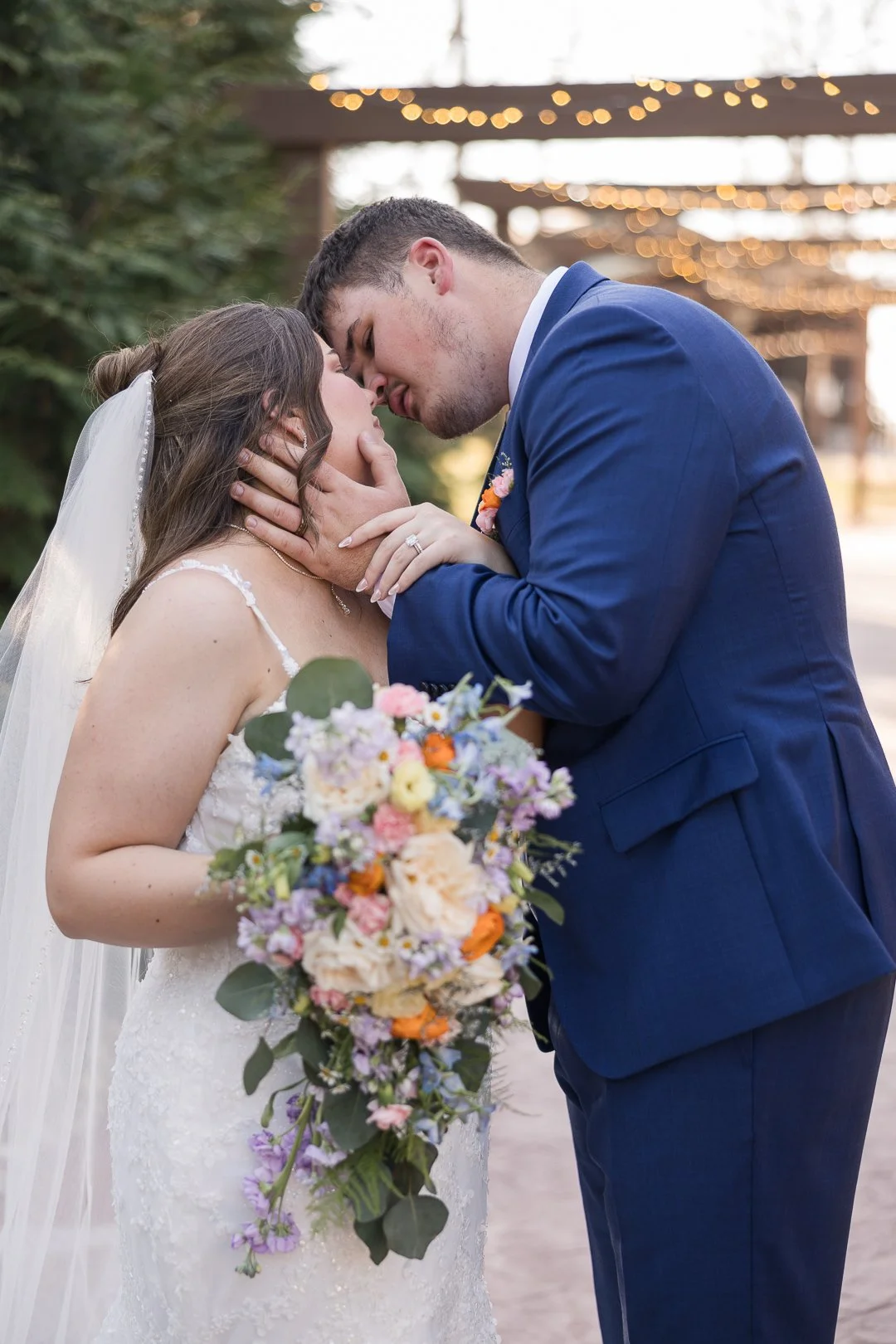 A bride and groom share a romantic moment at their wedding, with the groom gently holding the bride's face as they about to kiss. The bride holds a colorful bouquet of flowers, and the background features blurred string lights and outdoor setting.