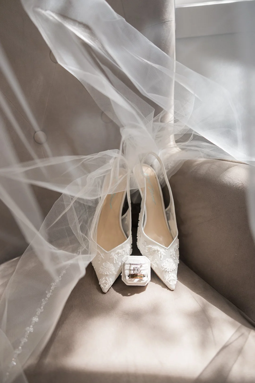 A pair of elegant white high-heeled wedding shoes with floral embellishments, placed on a cushioned surface with a wedding veil draped over them and an engagement ring in a box in front.
