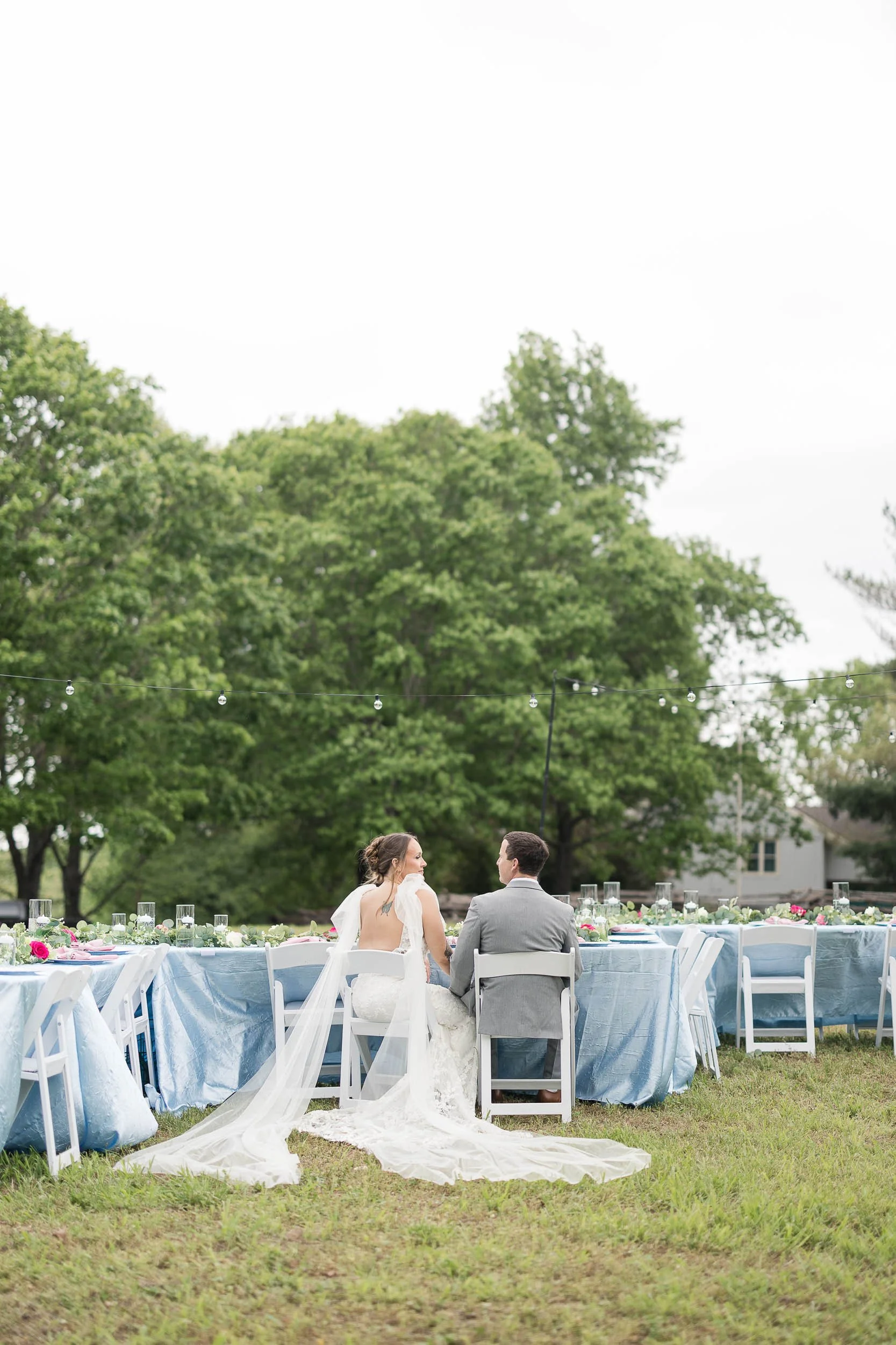 Bride and groom sitting at a decorated outdoor wedding reception table, facing each other, with lush green trees and string lights overhead.