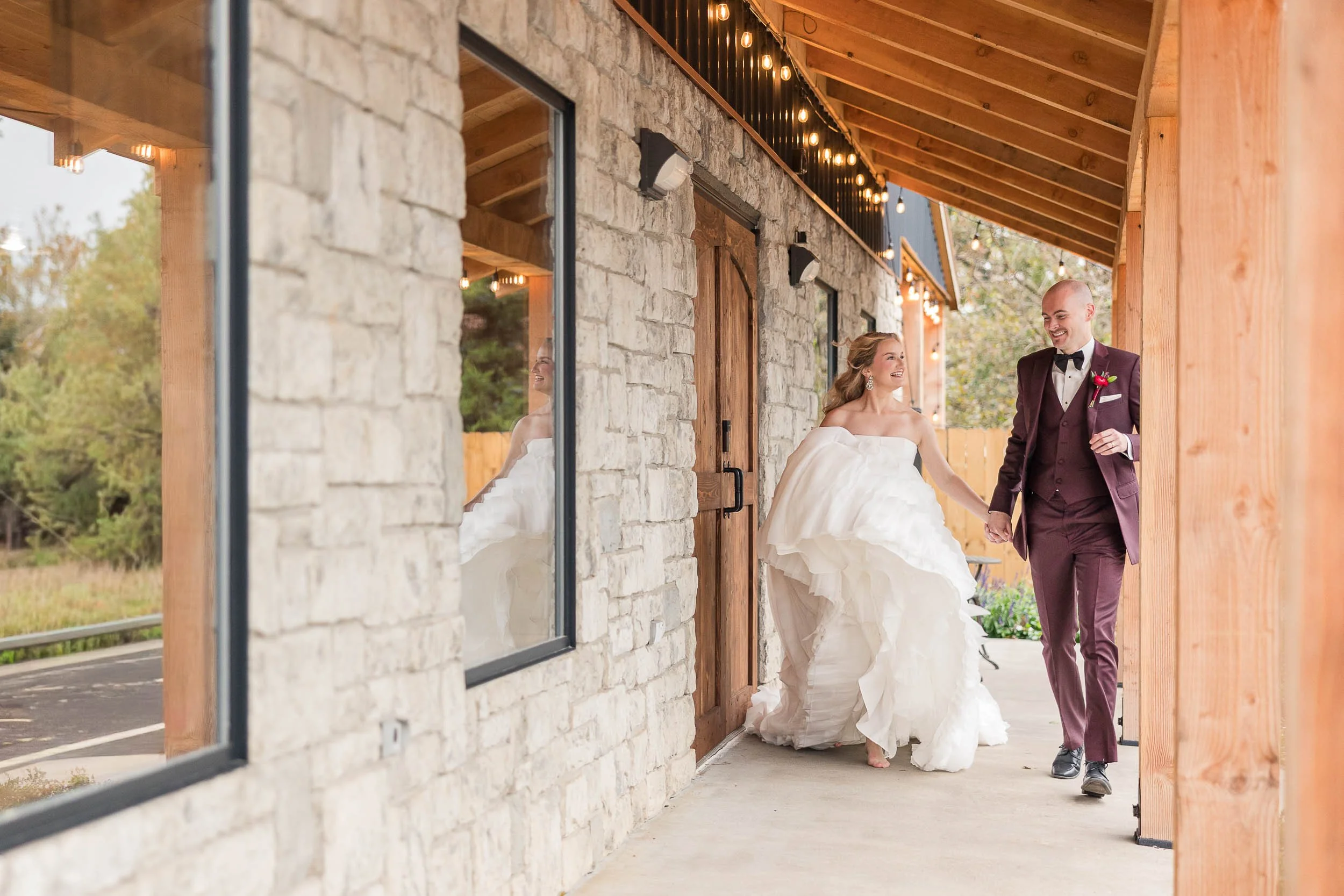 A bride and groom holding hands and smiling, walking outside a rustic building with stone walls and wooden accents, during a wedding celebration with string lights overhead.