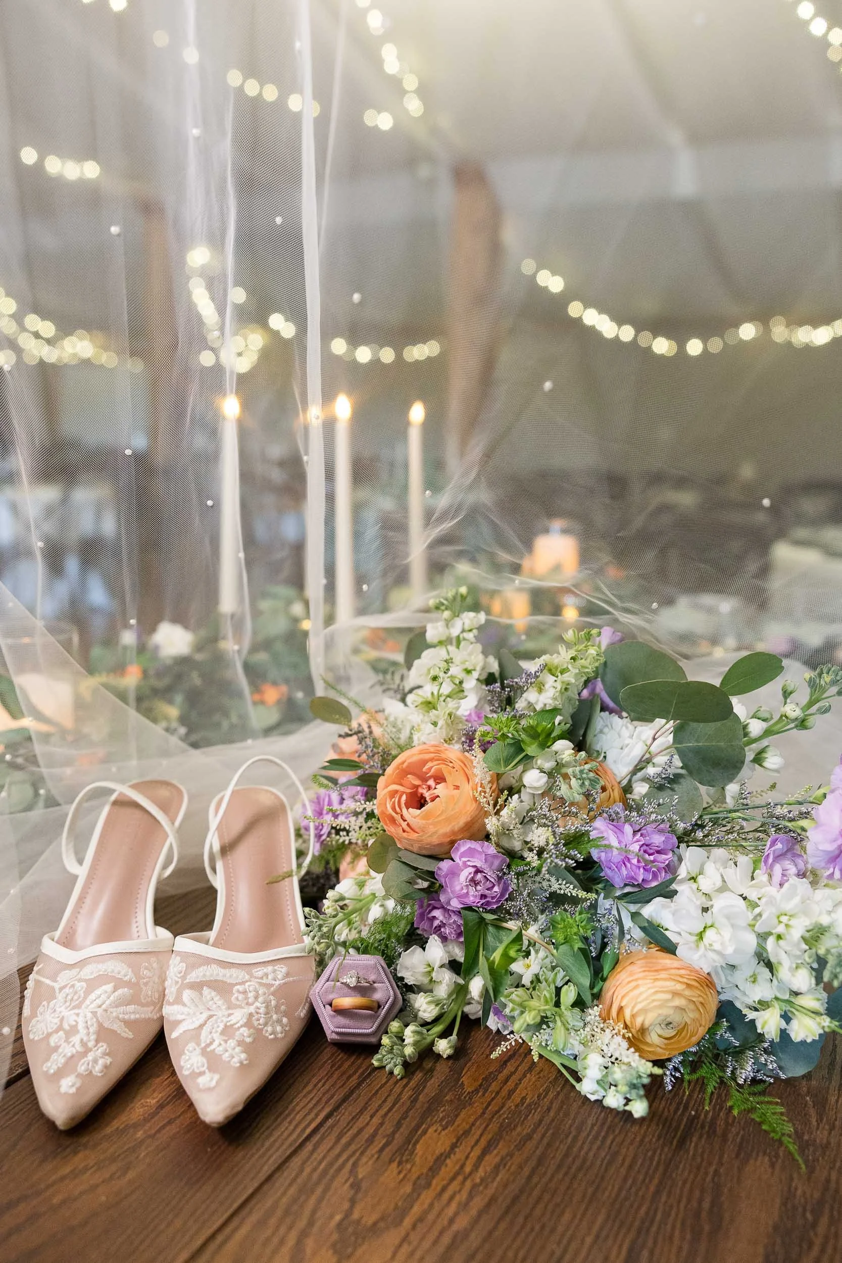 A bouquet of mixed flowers with orange roses, white and purple flowers, and greenery, placed on a wooden surface. Beside the bouquet are a pair of pink embroidered shoes with pointed toes, a small purple purse, and a pair of beige high heels. In the background, there are fairy lights and decorative star-shaped objects hanging from the ceiling.