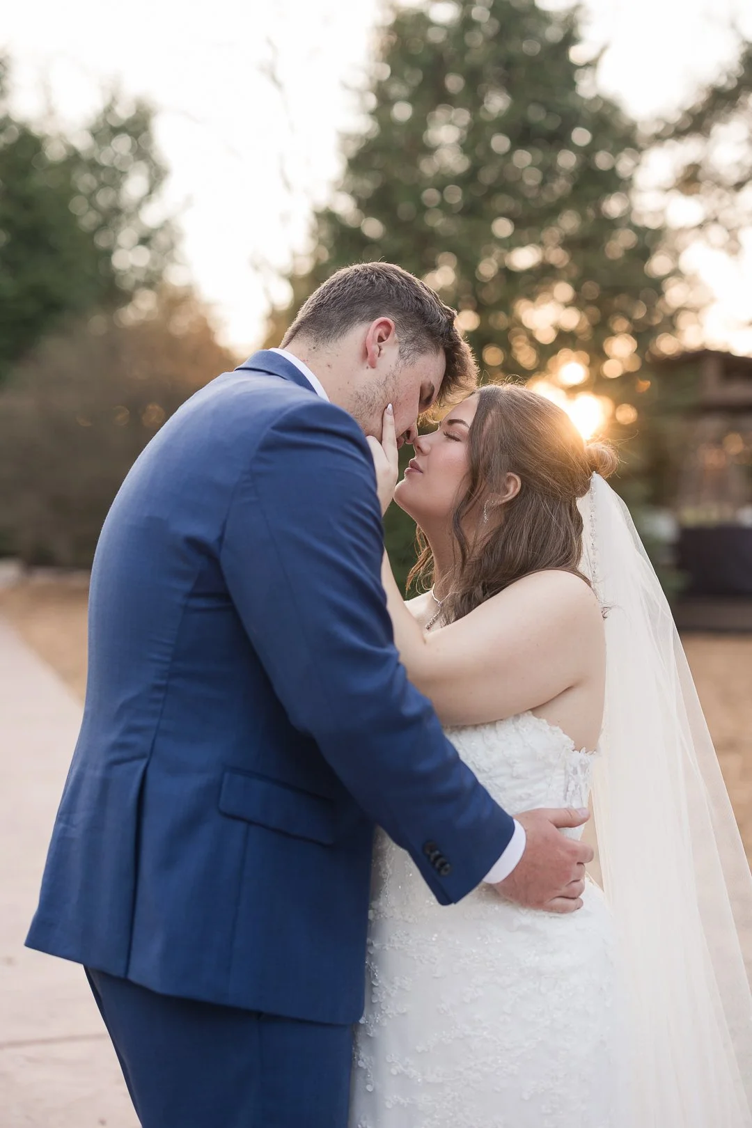 A bride and groom embrace outdoors during sunset, with their faces close together. The groom is dressed in a blue suit, and the bride is in a white wedding gown with a veil. They are sharing an intimate moment.