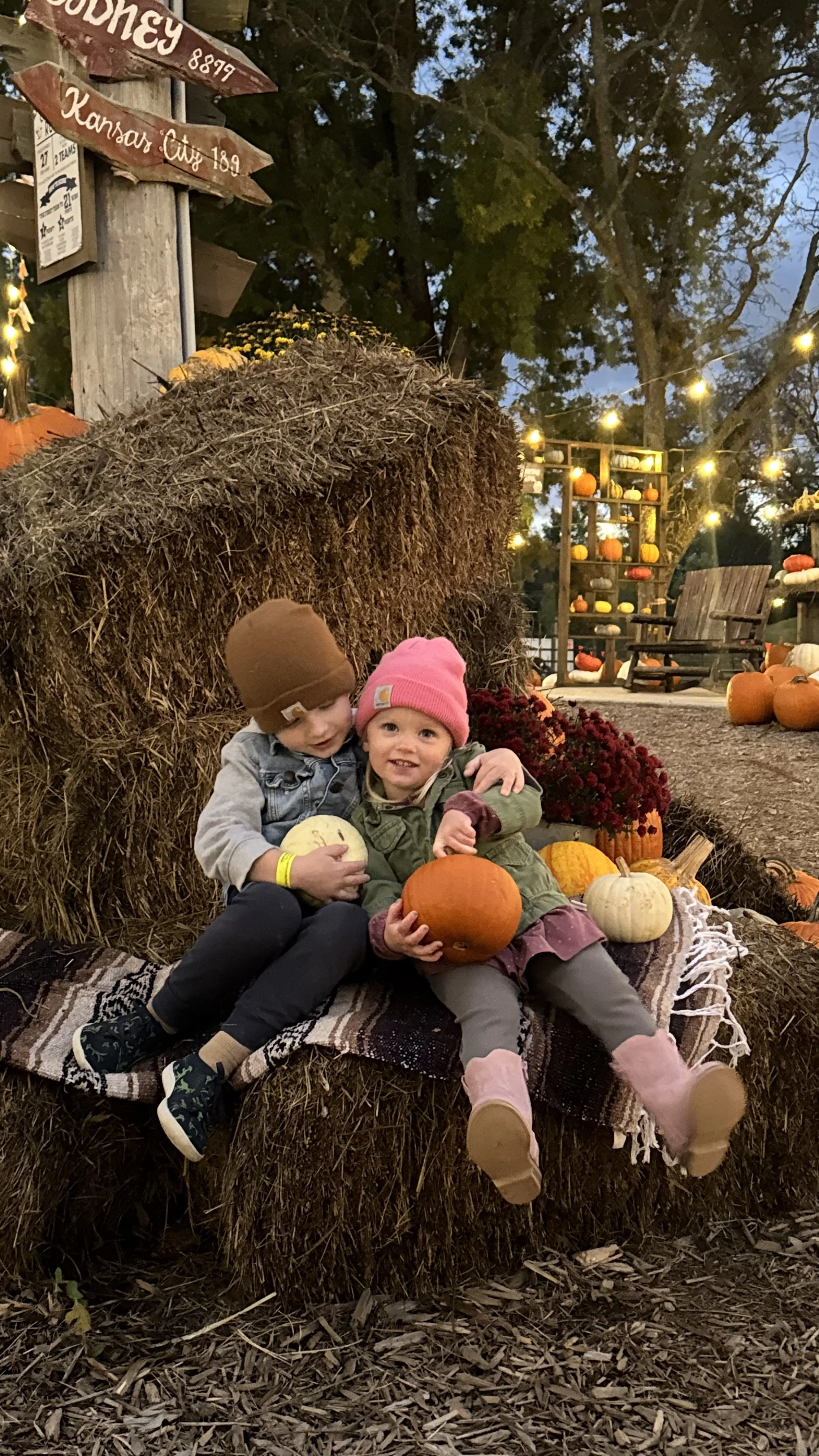 Two young children sitting on a hay bale at a fall pumpkin patch, holding pumpkins and surrounded by pumpkins and fall decorations, with string lights and trees in the background.