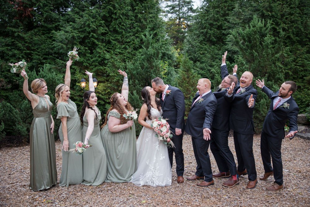Bride and groom surrounded by bridesmaids in green dresses and groomsmen in suits, posing playfully in an outdoor setting.