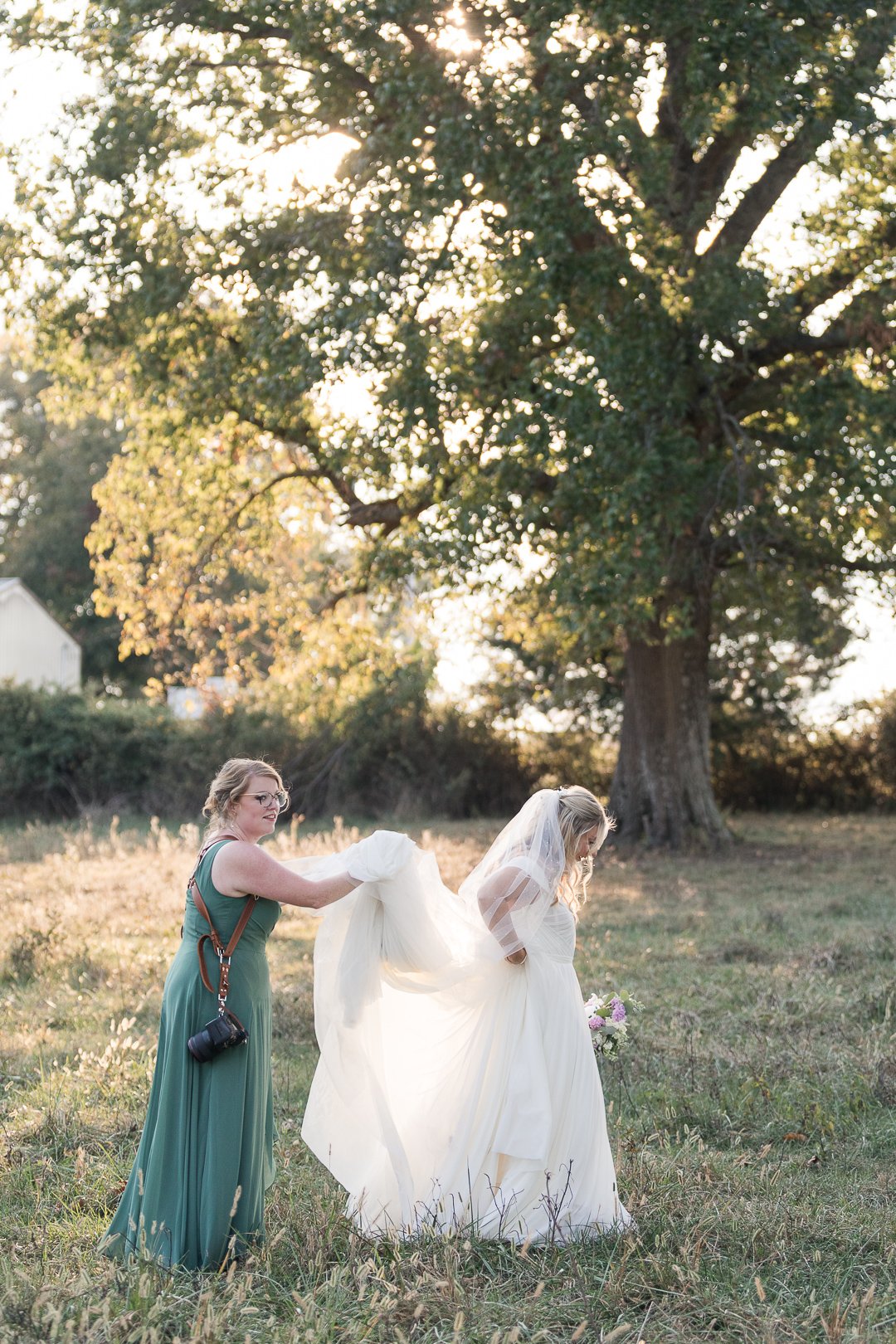 A bride in a white wedding dress holding a bouquet and a woman in a green dress assisting her outdoors near a large tree at sunset.