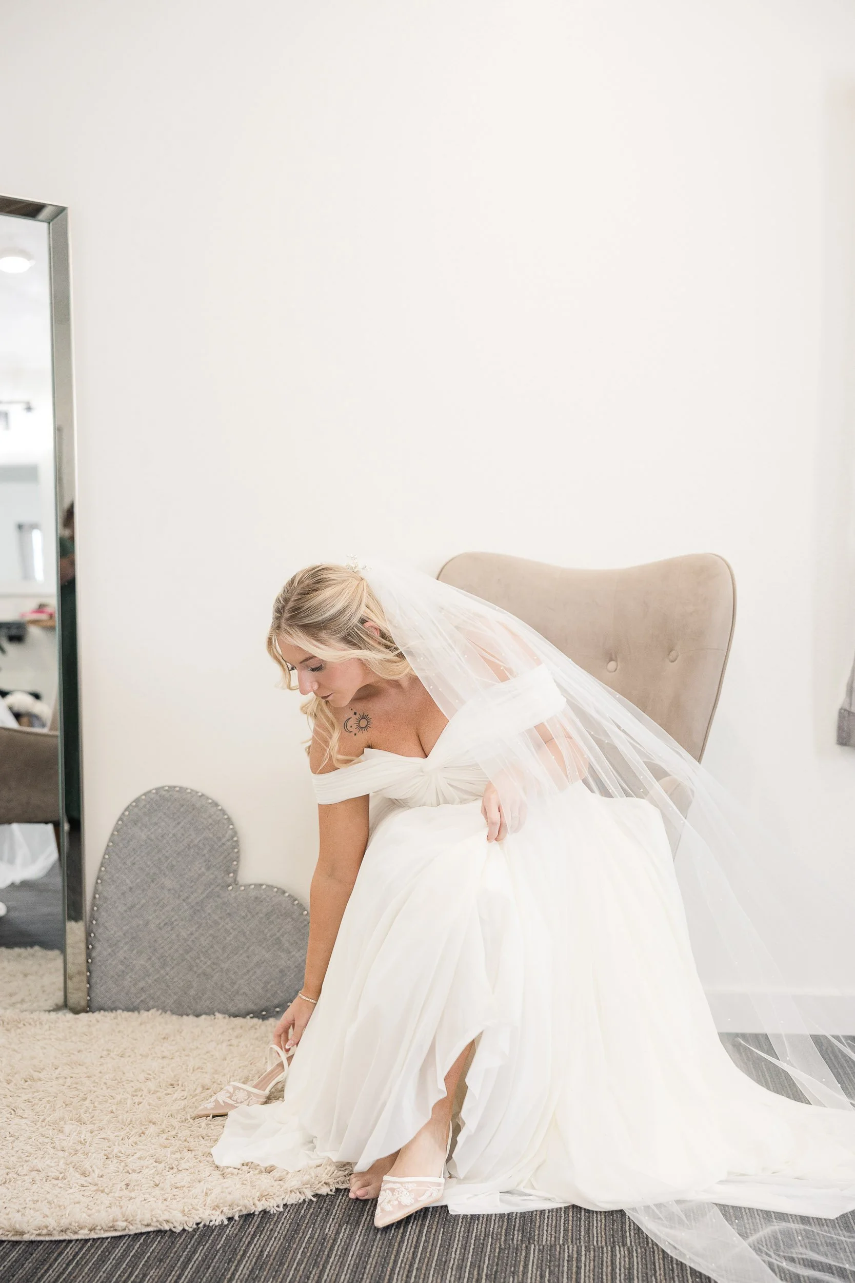 A bride in a white wedding dress and veil is bending down, adjusting her shoe in a room with cream-colored walls and a gray chair.