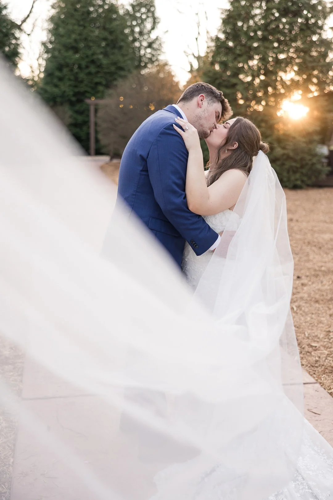 A bride and groom share a kiss outdoors during sunset, with trees in the background and the bride's veil flowing.