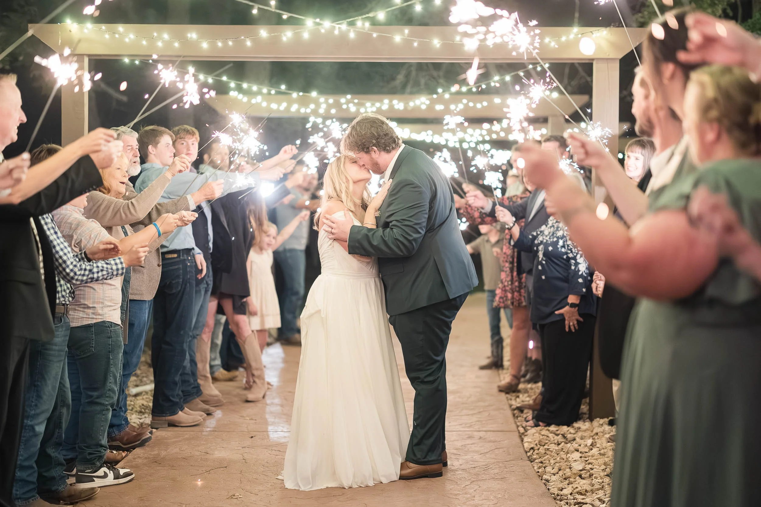 A couple in wedding attire sharing a kiss, surrounded by friends holding sparklers at an outdoor wedding reception at night.
