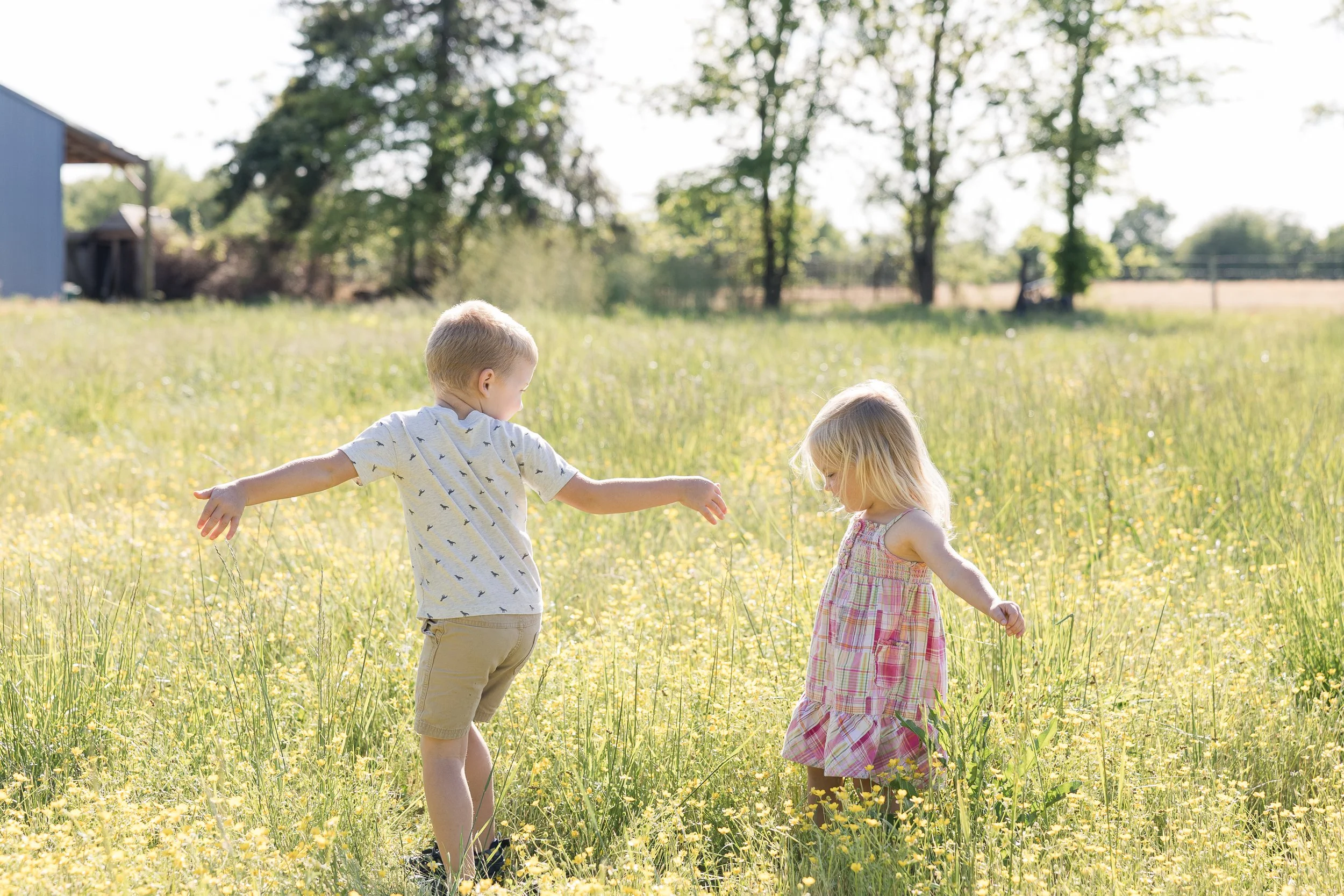 Two young children, a boy and a girl, holding hands and playing in a grassy, flower-filled field on a sunny day, with trees and a building in the background.